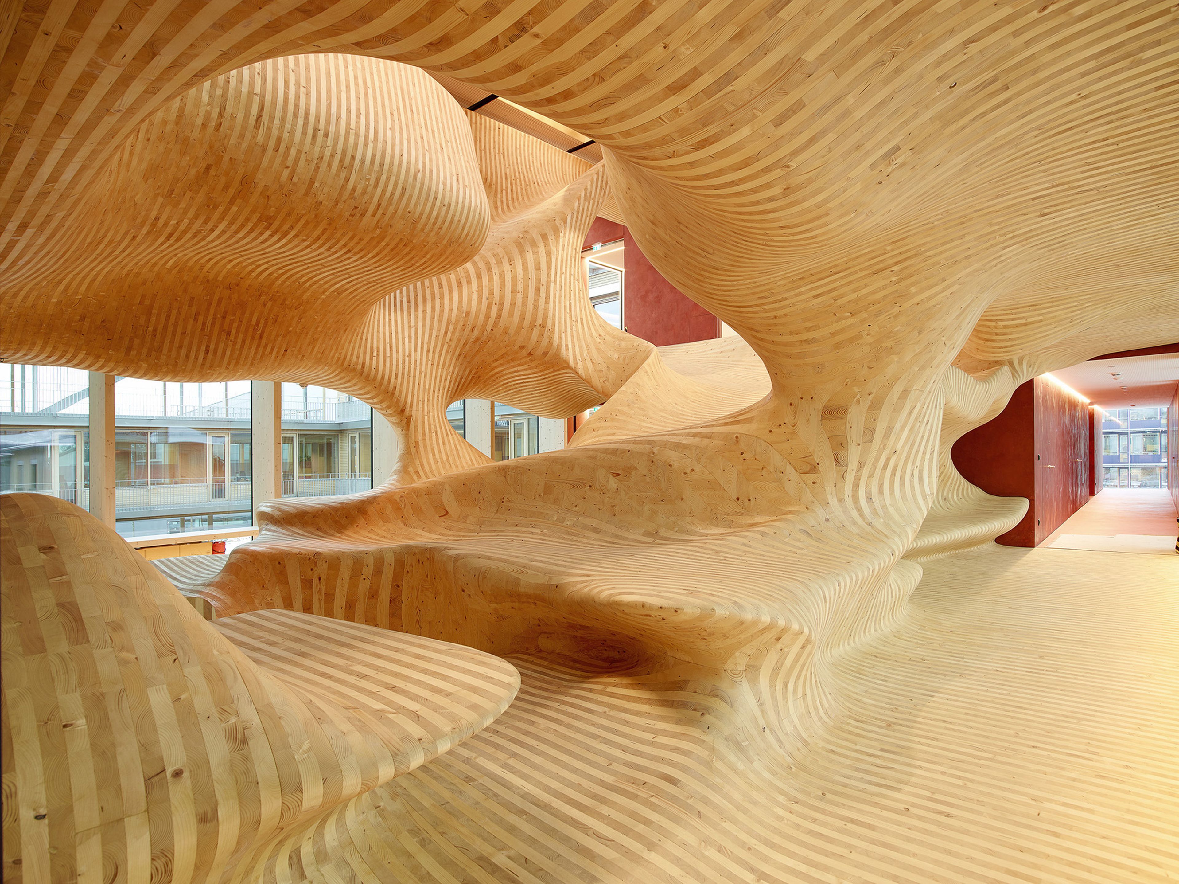 Modernes Interieur mit fließenden Holzstrukturen, die ein wellenförmiges Design erzeugen. Große Fenster sorgen für natürliches Licht und Ausblick.