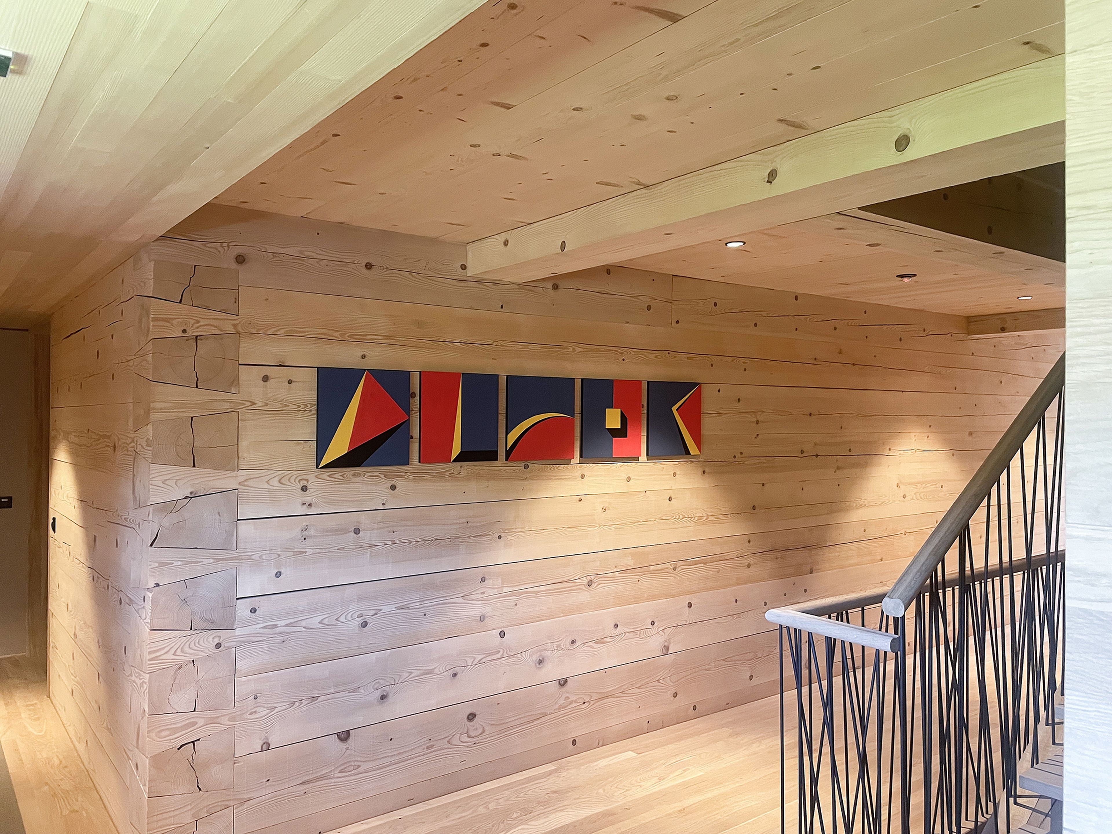 Holzvertäfelter Flur mit geometrischen Kunstwerken an der Wand und einem Geländer mit dunklen, vertikalen Stäben.