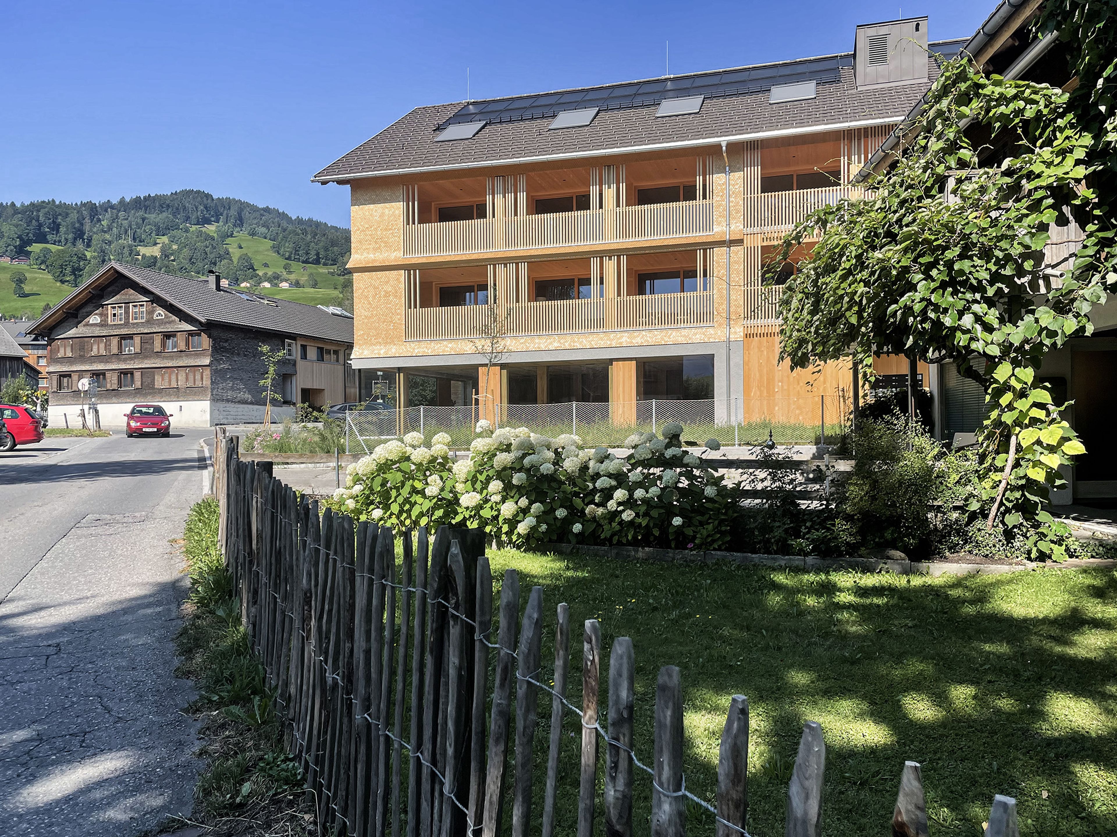 Modernes Holzgebäude mit Balkonen und sattgrünem Rasen. Im Hintergrund traditionelle Bregenzerwälder Häuser