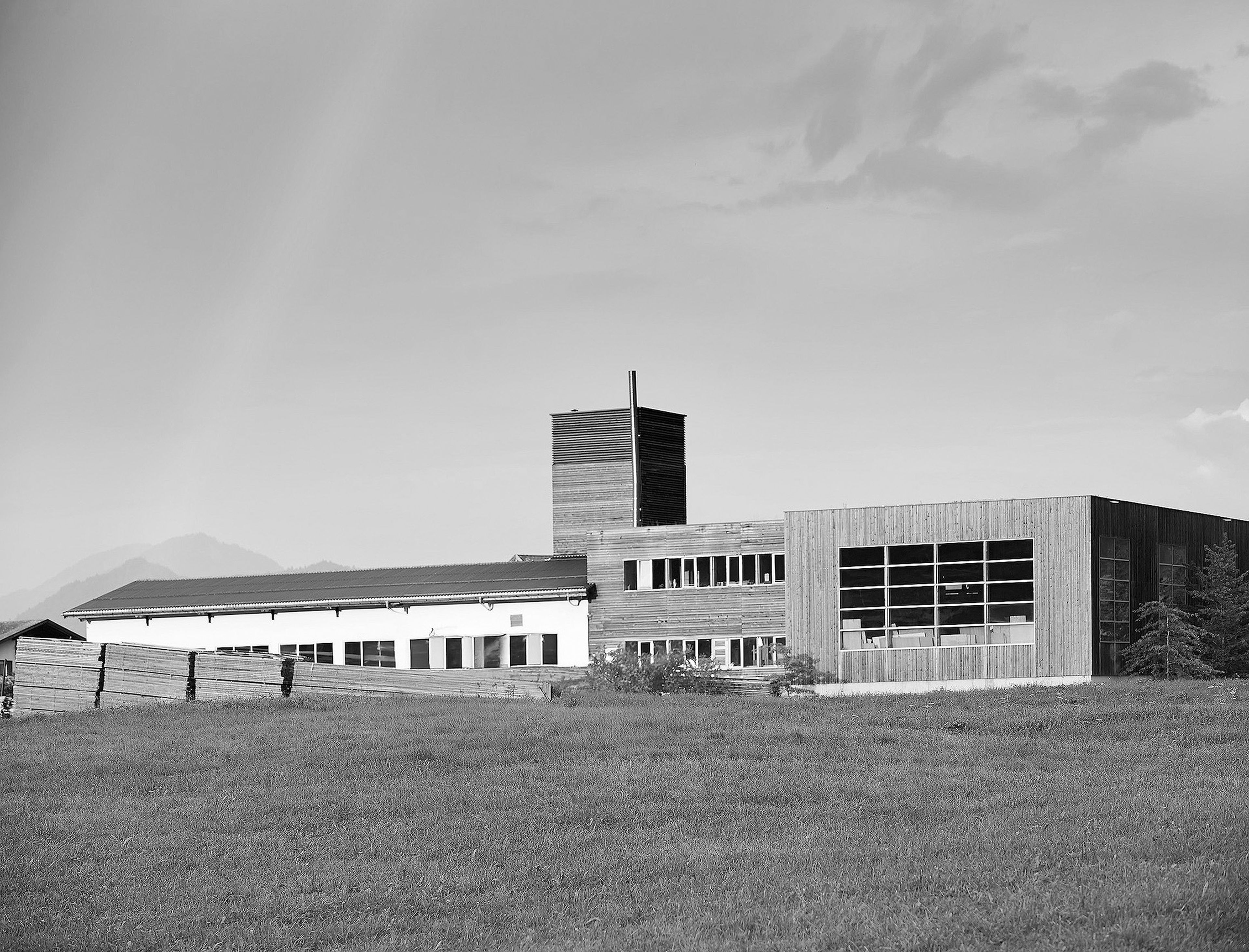 A large, modern industrial building with a rectangular wooden facade, set against a grassy field and a cloudy sky in grayscale.