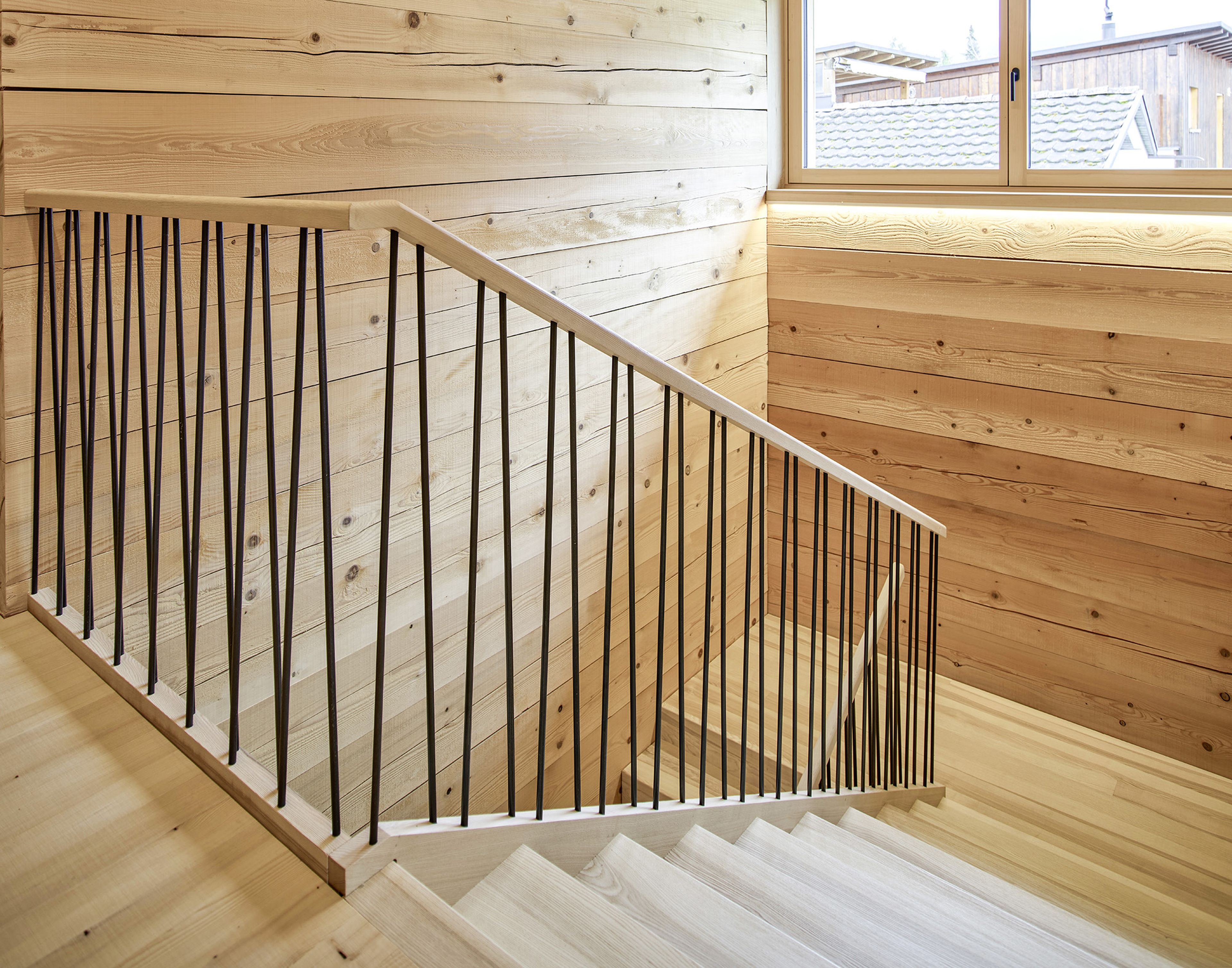 Eine Holztreppe mit schwarzem Metallgeländer, umgeben von hellen, holzgetäfelten Wänden, führt zu einem Fenster mit Blick auf die Dächer.