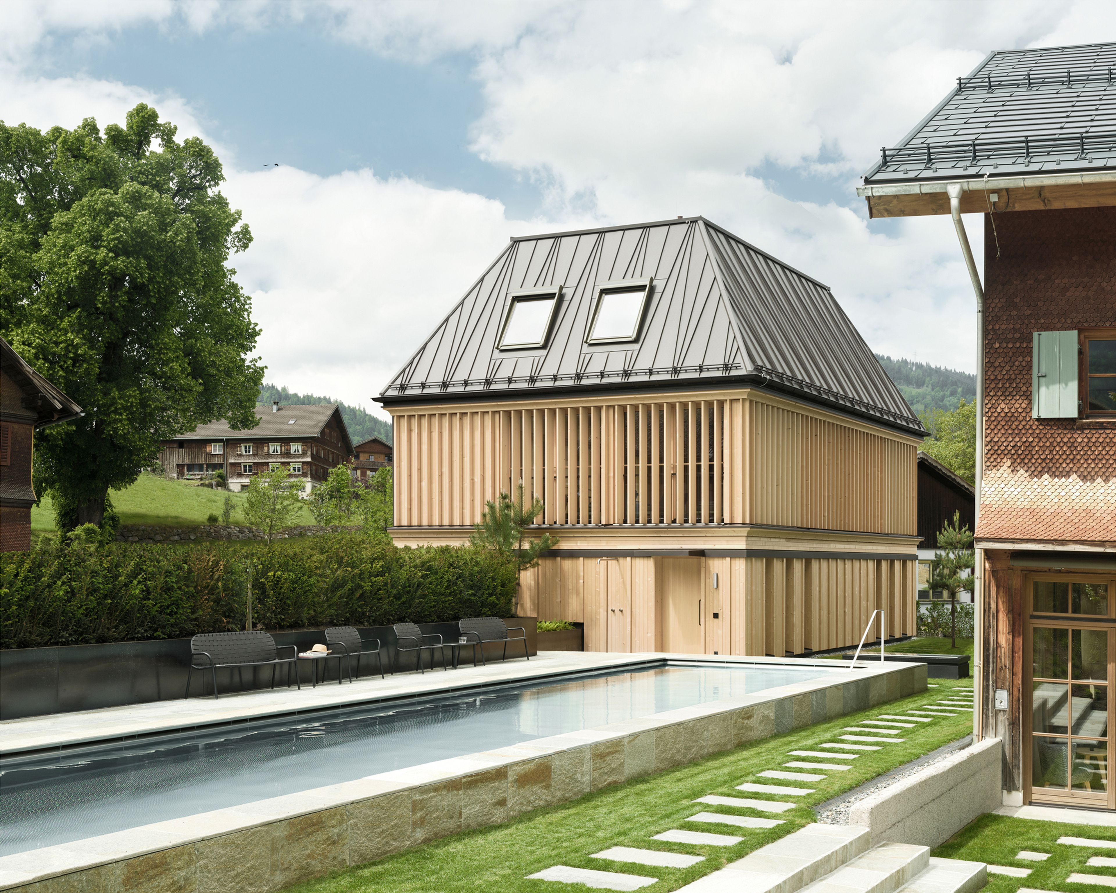 Modernes Holzhaus mit schrägem Metalldach, umgeben von Grün. Ein Steinweg führt zu einem Pool unter leicht bewölktem Himmel.