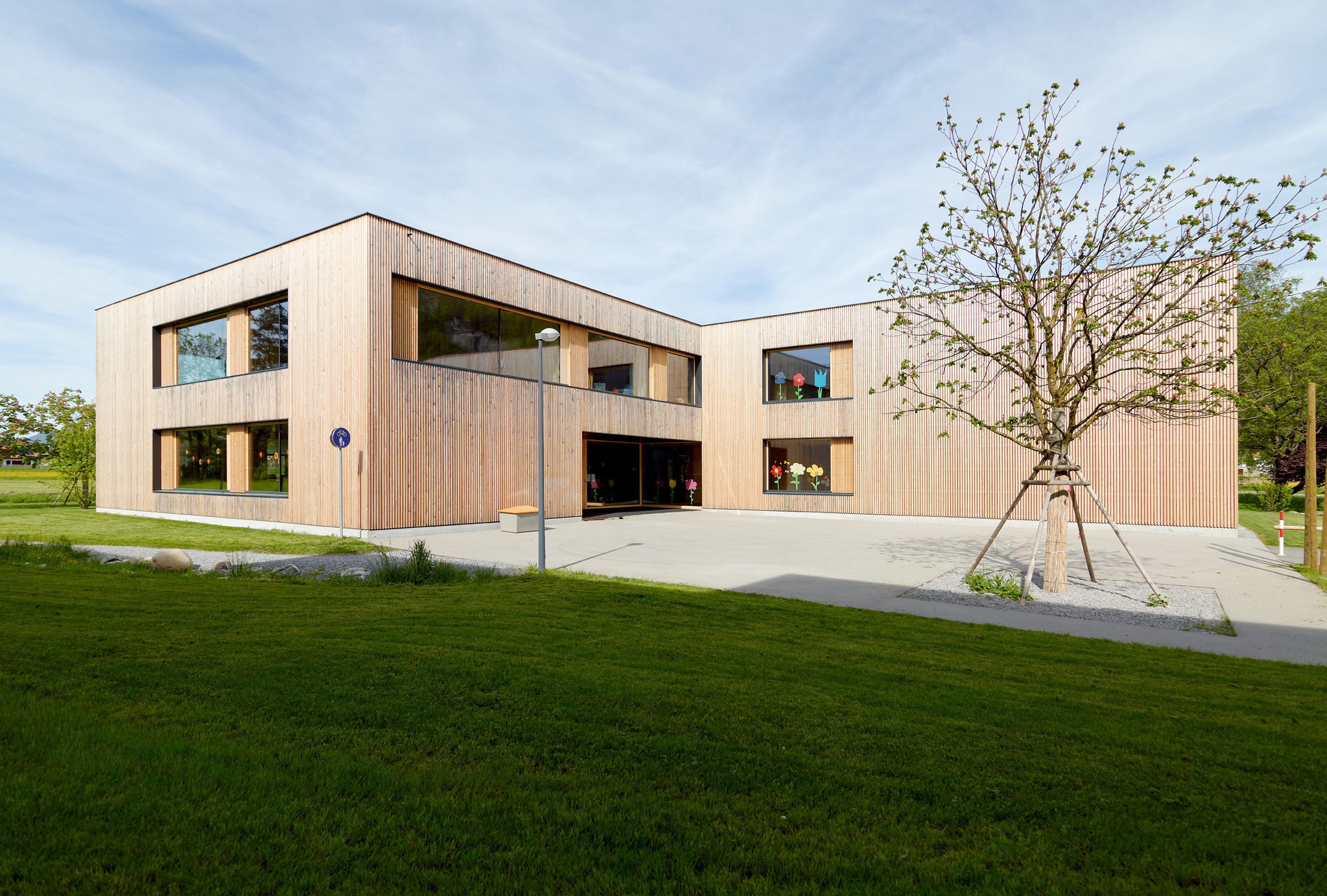 Moderner Kindergarten in nachhaltiger Holzbauweise mit klarer, zeitgemäßer Architektur, umgeben von viel Grün. Die natürliche Holzfassade fügt sich harmonisch in die Landschaft ein, große Fensterflächen sorgen für helle Räume und einen offenen Eindruck. Das Gebäude steht eingebettet zwischen Bäumen und Wiesen, mit naturnah gestalteten Außenbereichen und Spielzonen. Die warme Ausstrahlung des Holzes und die moderne Formensprache vermitteln Nachhaltigkeit, Geborgenheit und eine kinderfreundliche Atmosphäre.
