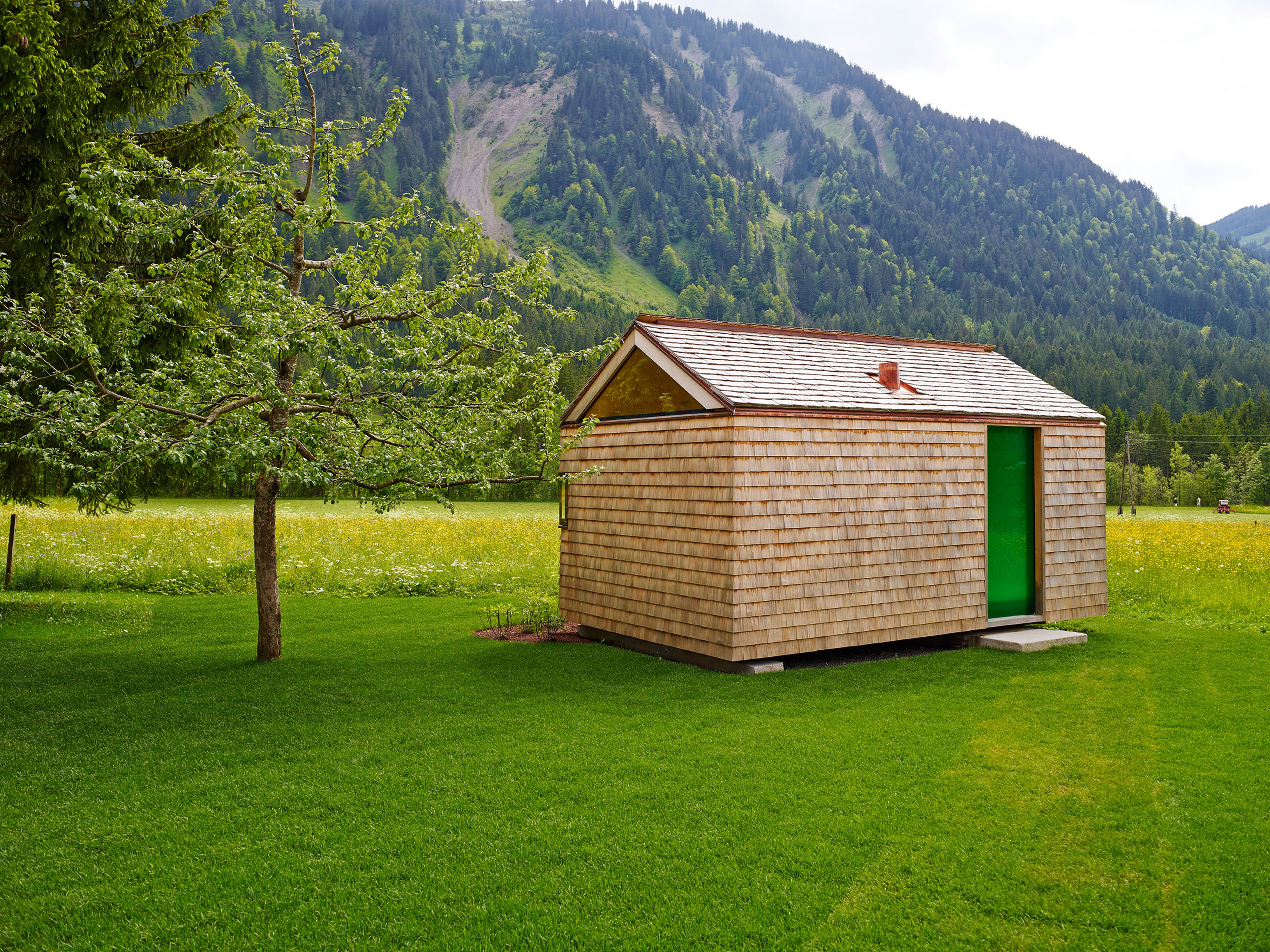 Kleines, modernes Gartenhaus in Holzbauweise mit Schindelfassade, klarer Formensprache und großem Fenster, umgeben von Grün.