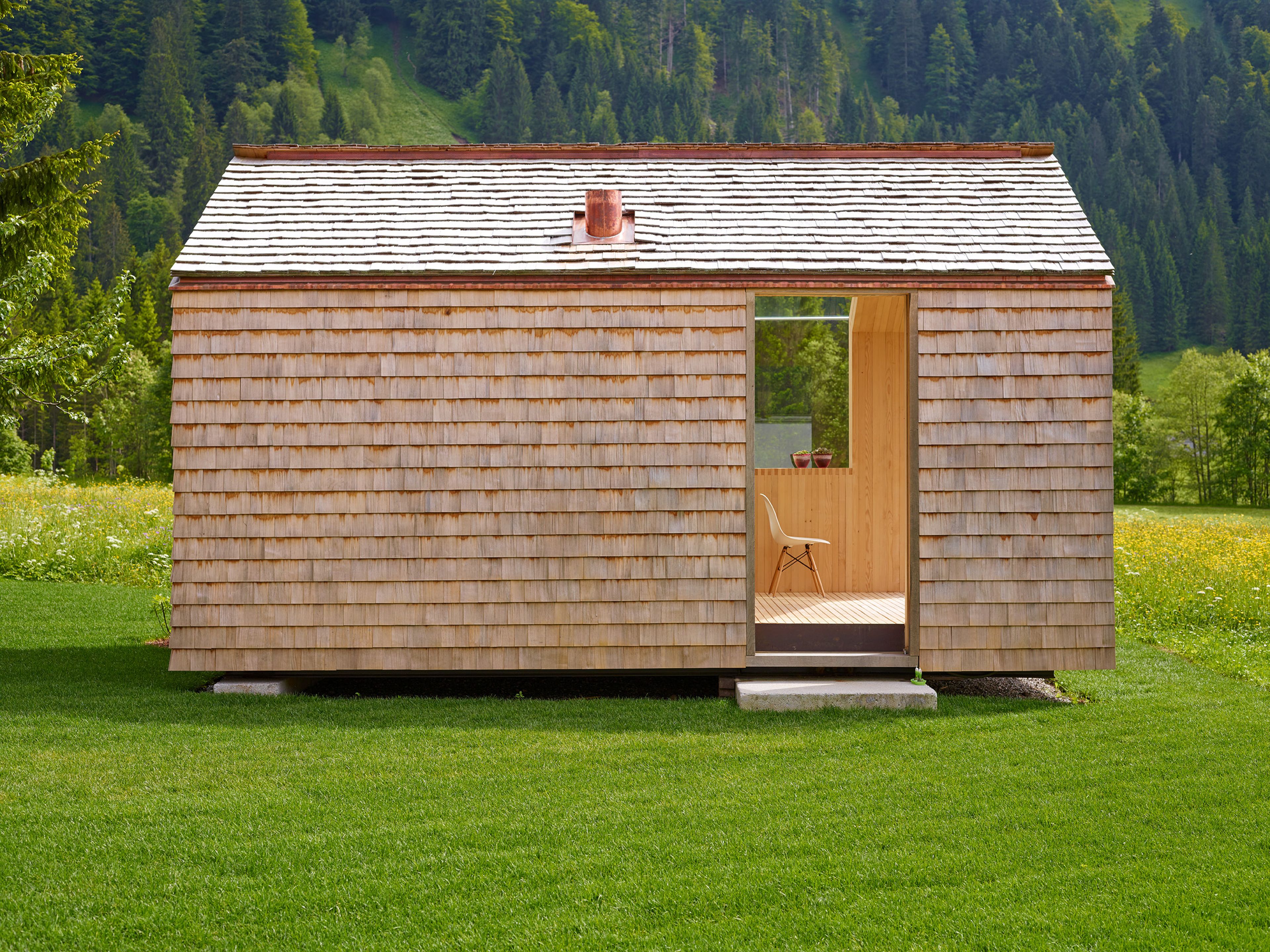 Kleines, modernes Gartenhaus in Holzbauweise mit Schindelfassade, klarer Formensprache und großem Fenster, umgeben von Grün.