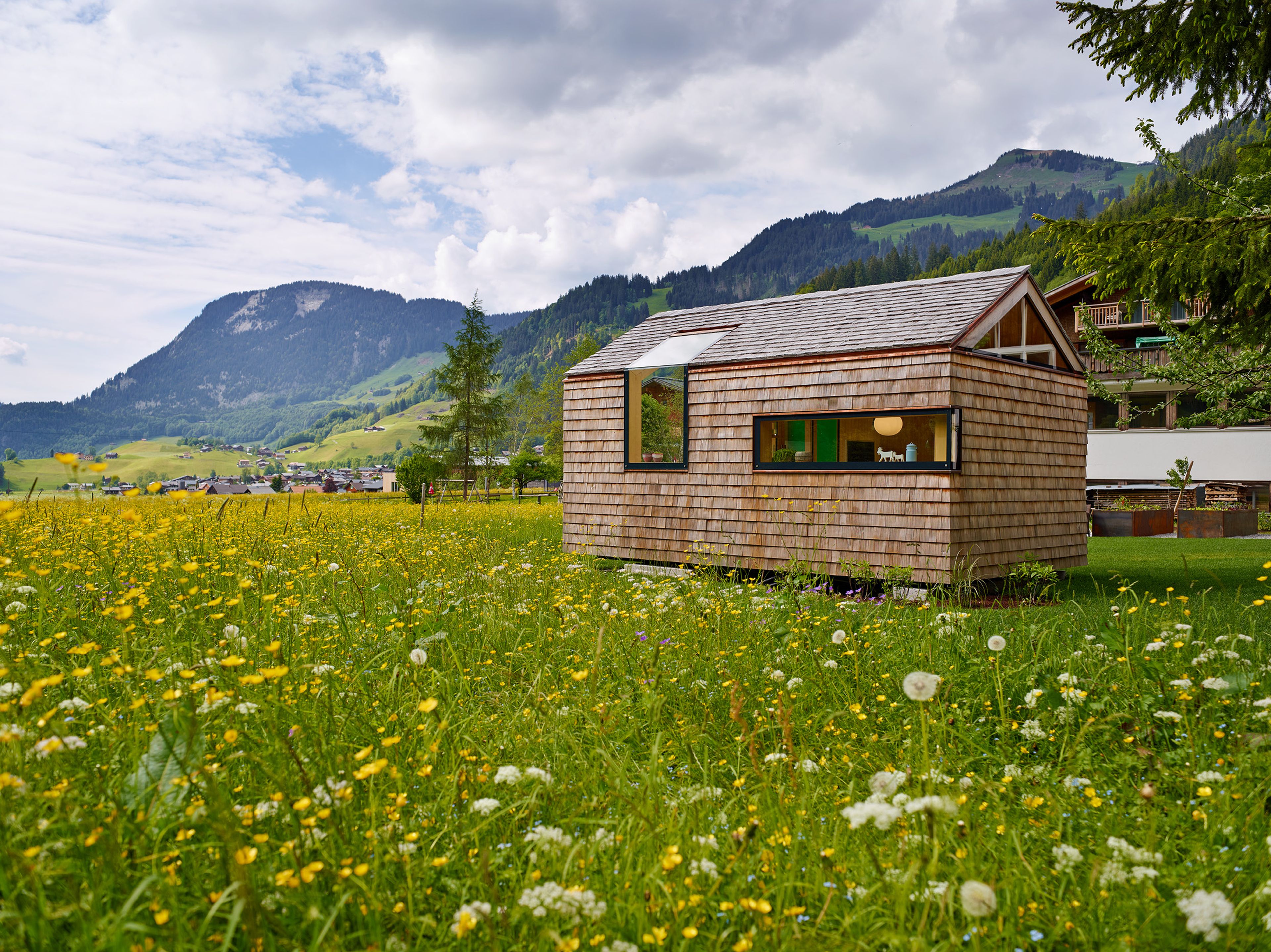 Kleines, modernes Gartenhaus in Holzbauweise mit Schindelfassade, klarer Formensprache und großem Fenster, umgeben von Grün.