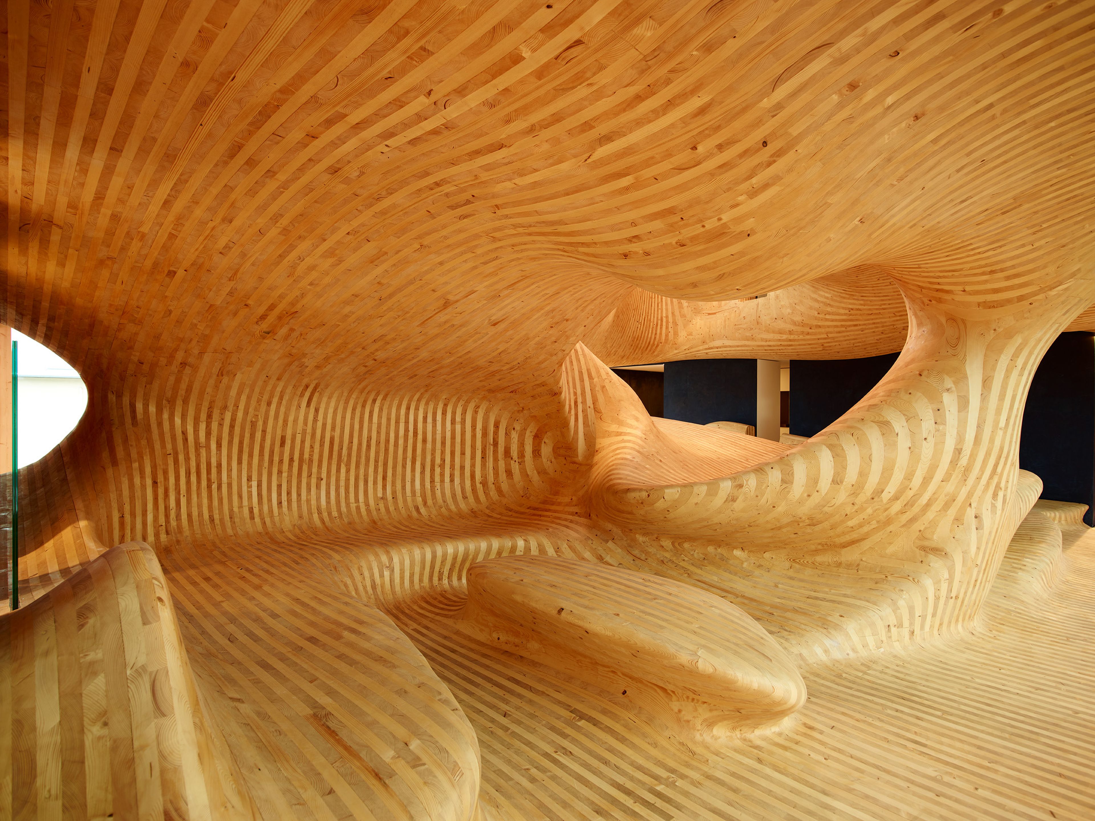 Begehbare, organisch geformte Holzskulptur ‚Body‘ im Innenraum des OMICRON-Gebäudes in Klaus, entworfen von Eichinger Offices und ausgeführt von Gerhard Berchtold Zimmerei – eine skulpturale Kommunikationszone aus laminierten Brettsperrholzflächen, die fließende, körperhafte Formen und kommunikative Begegnungsflächen erzeugt.