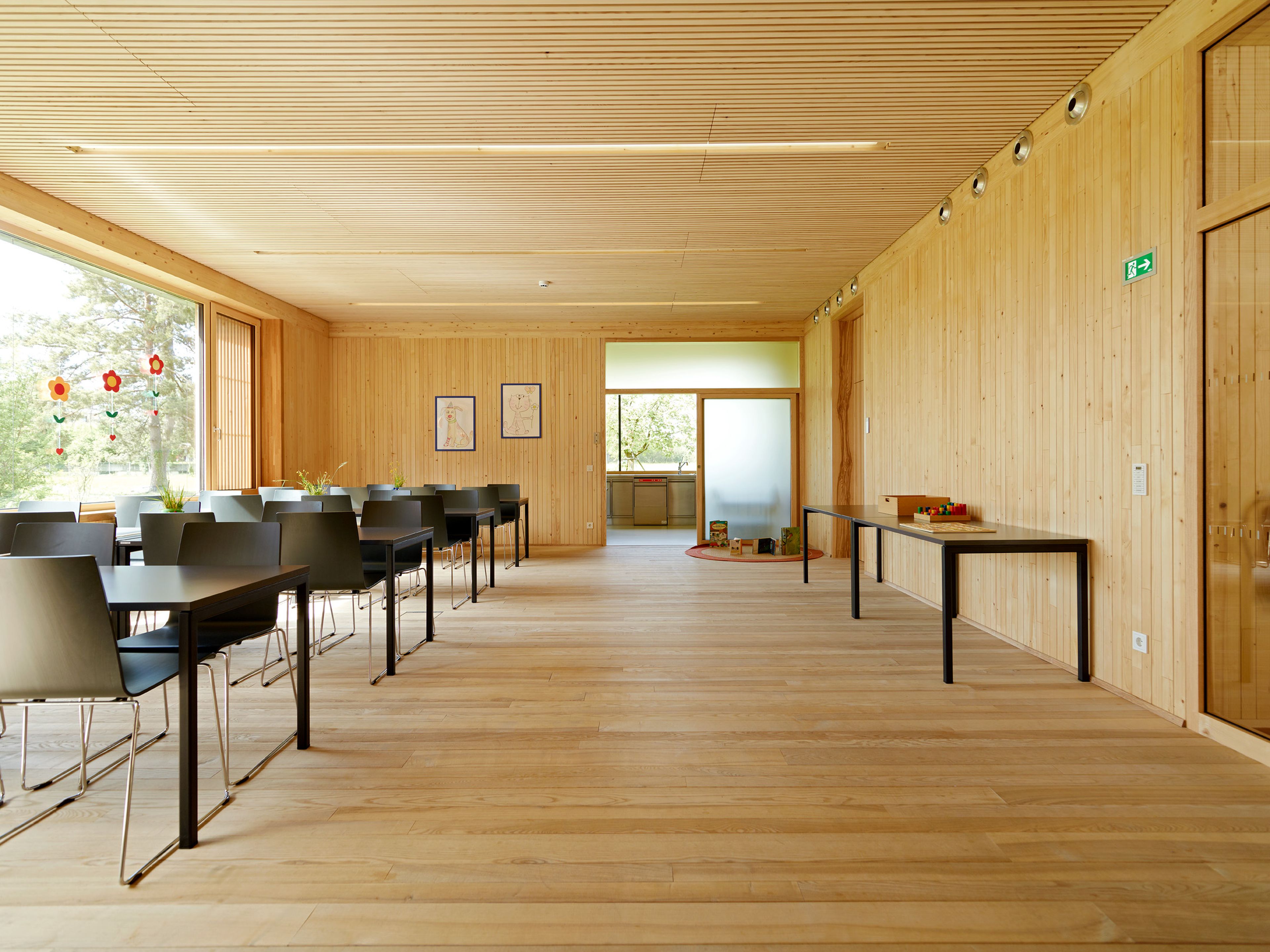 Innenraum eines Kindergartens mit durchgehendem Holzboden, Holzdecken und Holzwänden in heller, freundlicher Gestaltung. Die natürliche Materialität schafft ein gesundes Raumklima und eine warme, behagliche Umgebung. Lichtdurchflutete Räume mit klarer Struktur und kindgerechter Ausstattung laden zum Spielen, Lernen und Wohlfühlen ein. Die nachhaltige Holzbauweise vermittelt Sicherheit, Natürlichkeit und eine perfekte Umgebung für Kinder.