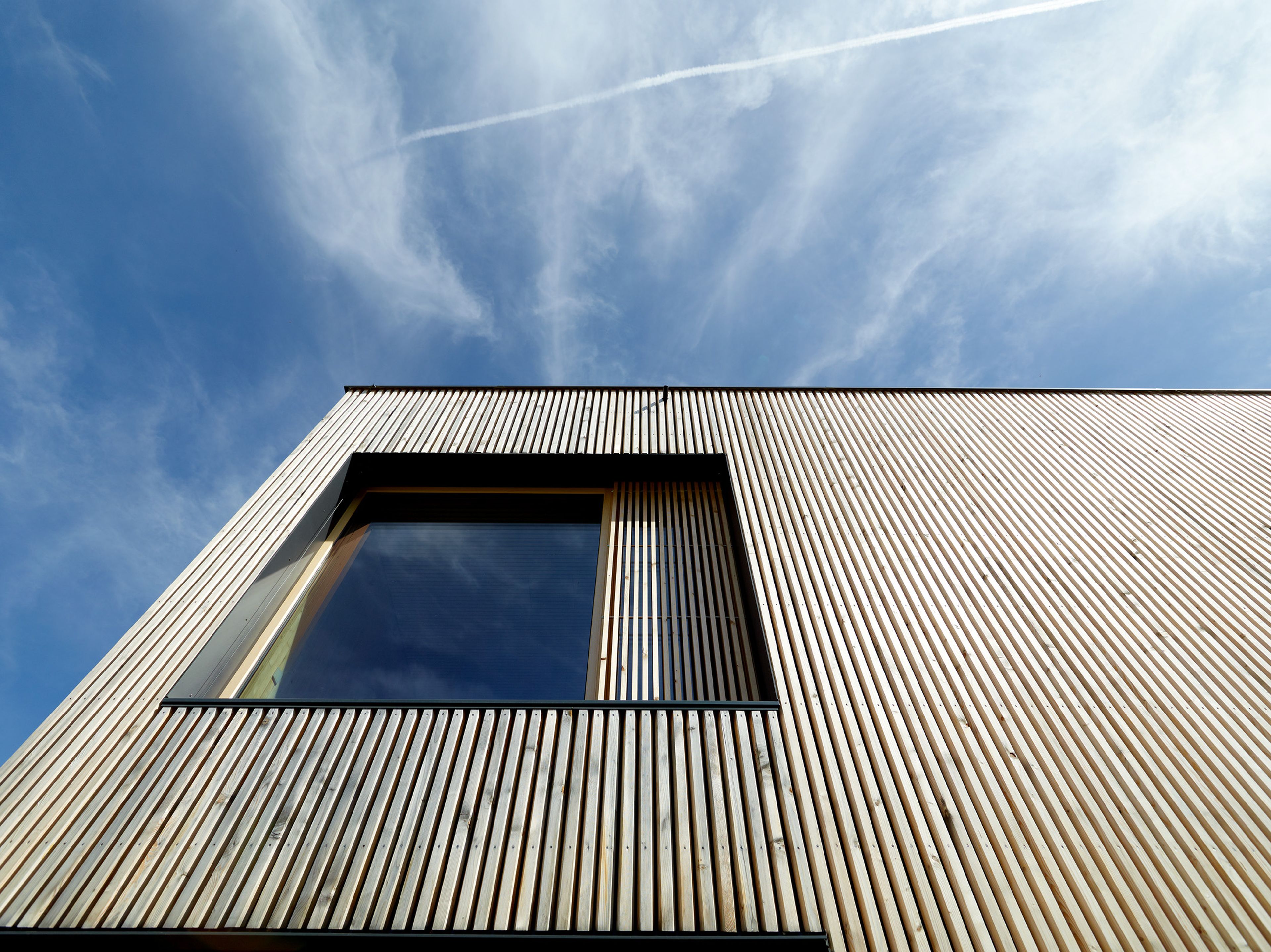 Detailaufnahme einer vertikal stehenden Holzfassade aus naturbelassenem Holz mit klarer Linienführung. Die senkrechten Holzlamellen zeigen natürliche Maserung und warme Farbtöne und unterstreichen die hochwertige, moderne Architektur des Gebäudes.