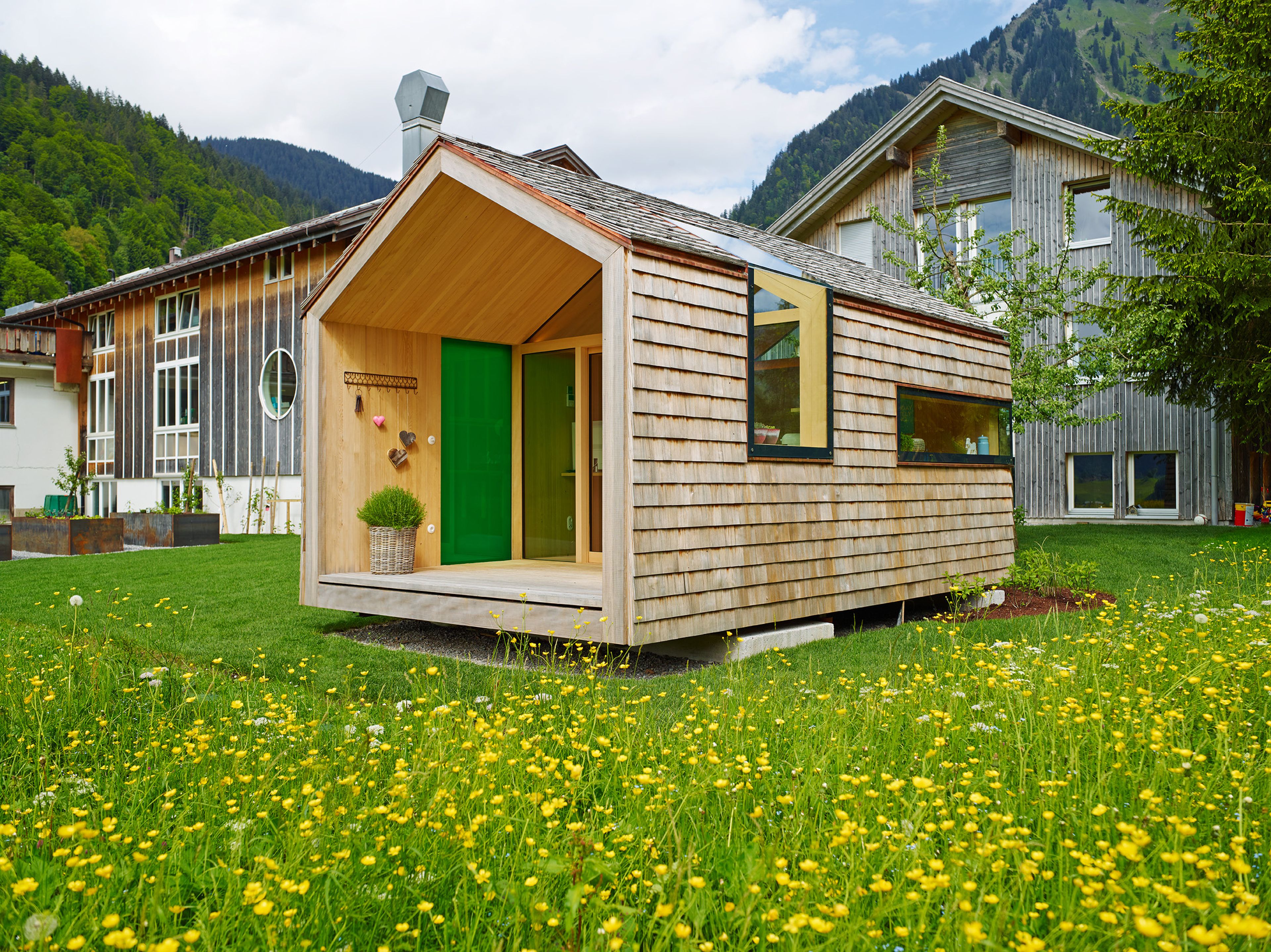 Kleines, modernes Gartenhaus in Holzbauweise mit Schindelfassade, klarer Formensprache und großem Fenster, umgeben von Grün.