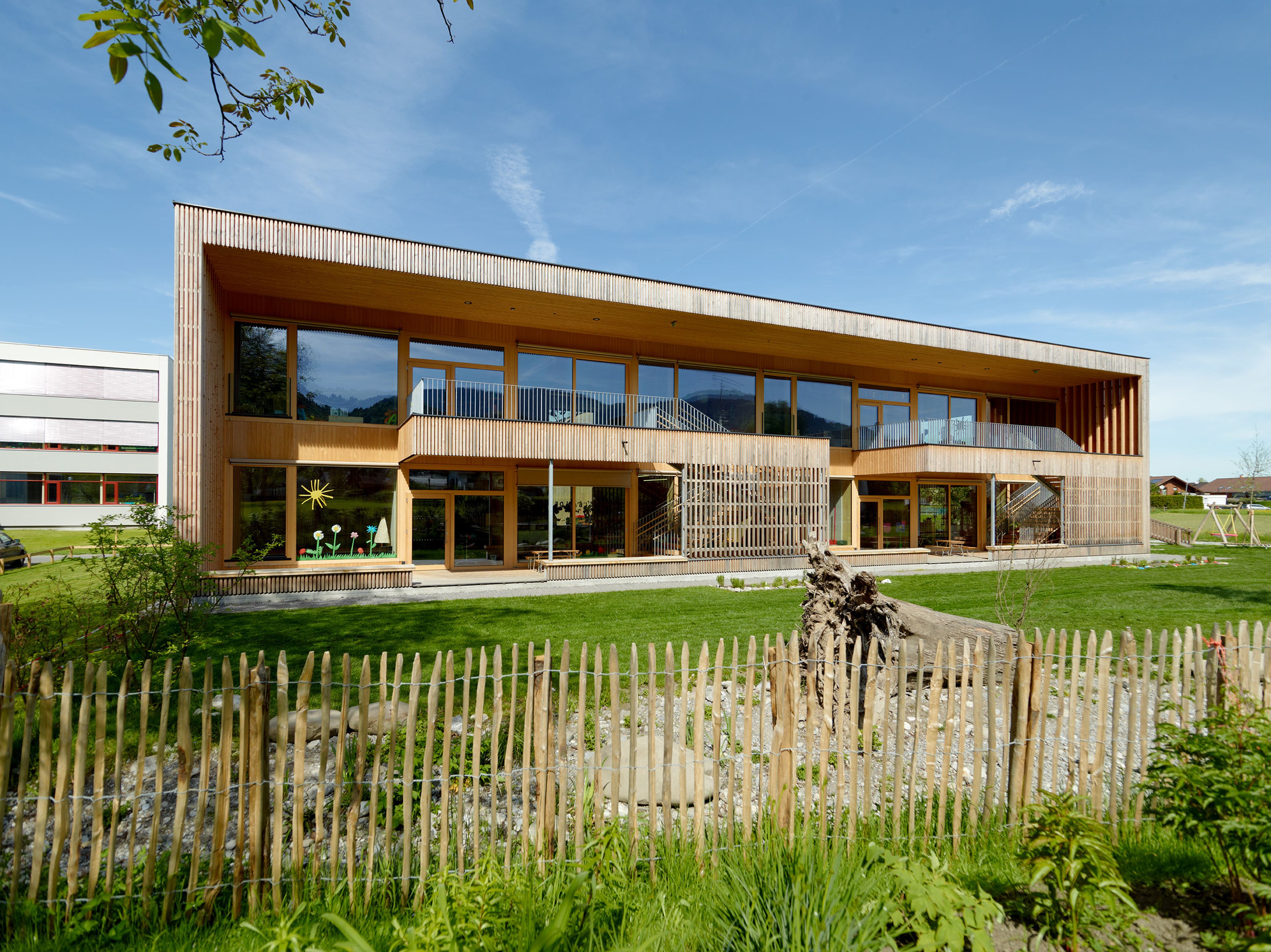 Moderner Kindergarten in nachhaltiger Holzbauweise mit klarer, zeitgemäßer Architektur, umgeben von viel Grün. Die natürliche Holzfassade fügt sich harmonisch in die Landschaft ein, große Fensterflächen sorgen für helle Räume und einen offenen Eindruck. Das Gebäude steht eingebettet zwischen Bäumen und Wiesen, mit naturnah gestalteten Außenbereichen und Spielzonen. Die warme Ausstrahlung des Holzes und die moderne Formensprache vermitteln Nachhaltigkeit, Geborgenheit und eine kinderfreundliche Atmosphäre.