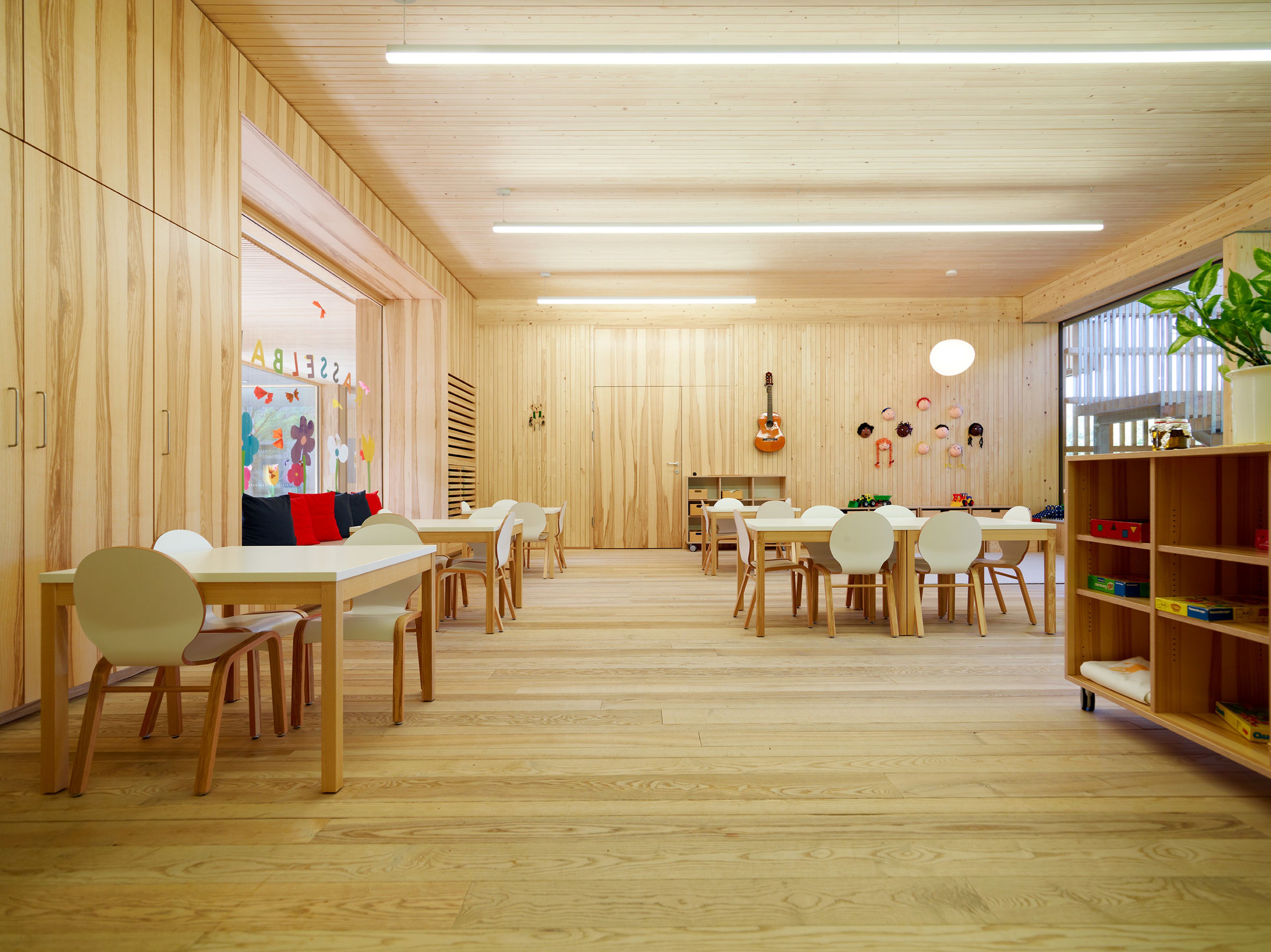 Innenraum eines Kindergartens mit durchgehendem Holzboden, Holzdecken und Holzwänden in heller, freundlicher Gestaltung. Die natürliche Materialität schafft ein gesundes Raumklima und eine warme, behagliche Umgebung. Lichtdurchflutete Räume mit klarer Struktur und kindgerechter Ausstattung laden zum Spielen, Lernen und Wohlfühlen ein. Die nachhaltige Holzbauweise vermittelt Sicherheit, Natürlichkeit und eine perfekte Umgebung für Kinder.