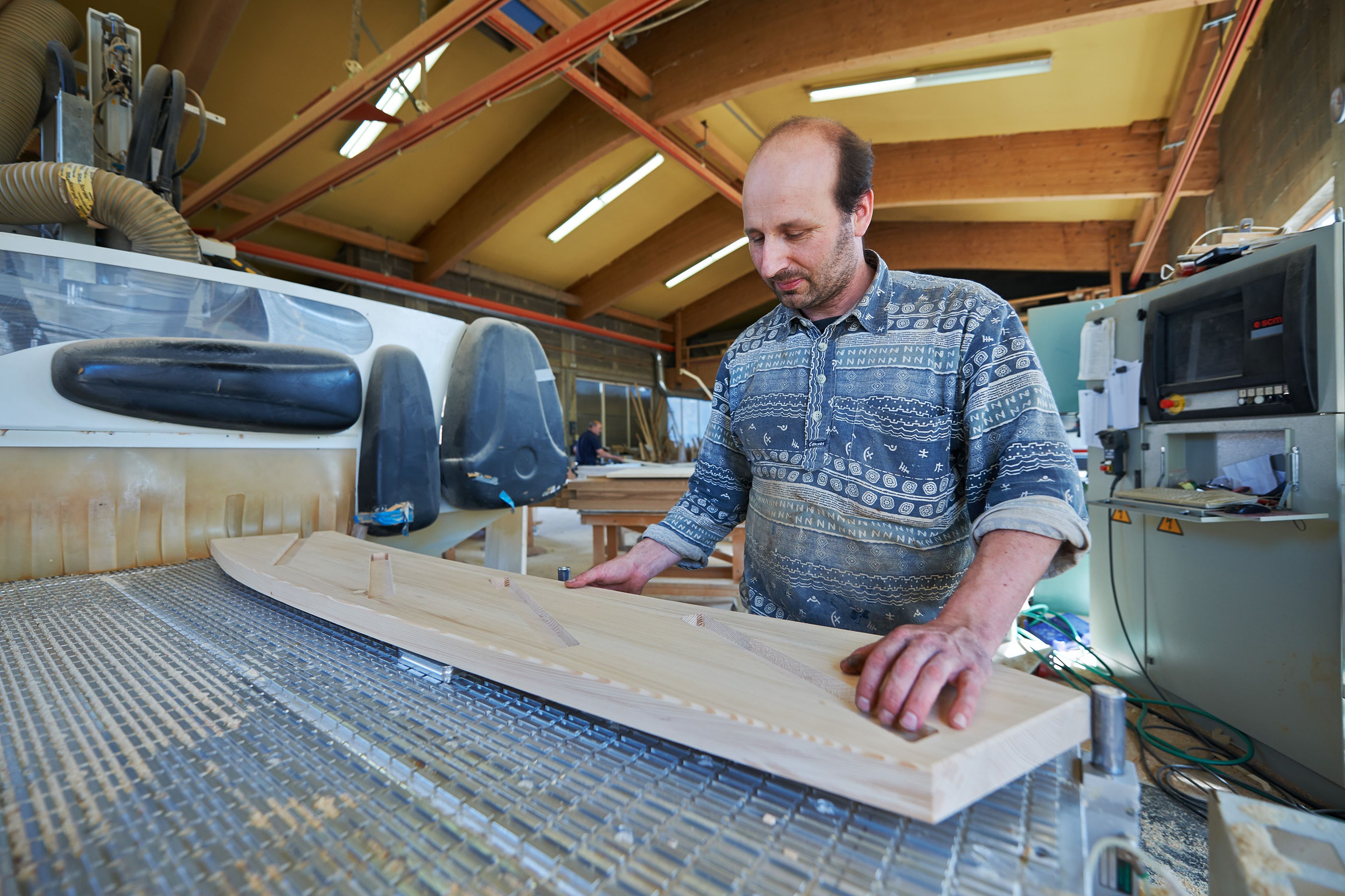 A man in a workshop uses a CNC machine to shape a wooden piece. The space is well-lit, with woodworking tools visible in the background.