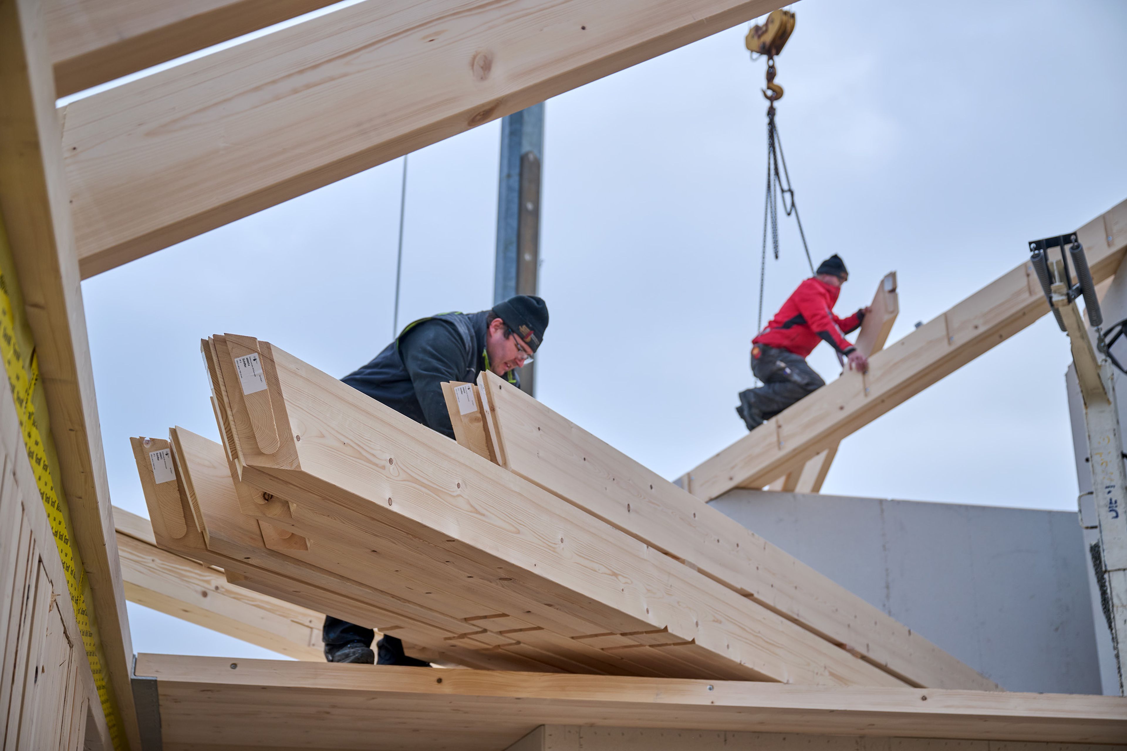Zwei Zimmerer montieren Holzbalken an einer Konstruktion; einer von ihnen steht auf einem Gerüst, im Hintergrund ist ein Kran zu sehen.