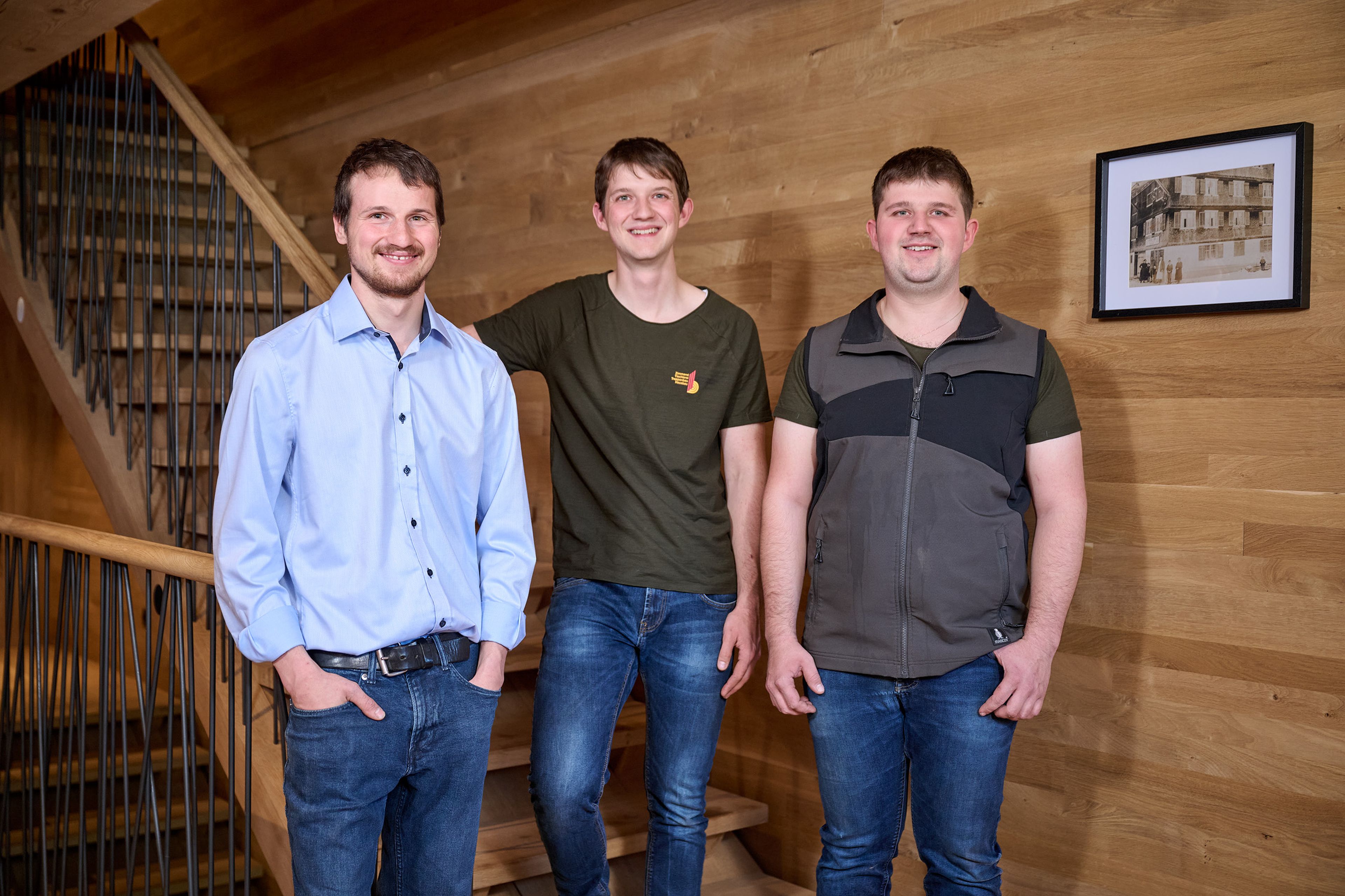 Drei Männer stehen lächelnd und lässig in Jeans und Hemden gekleidet auf einer Holztreppe