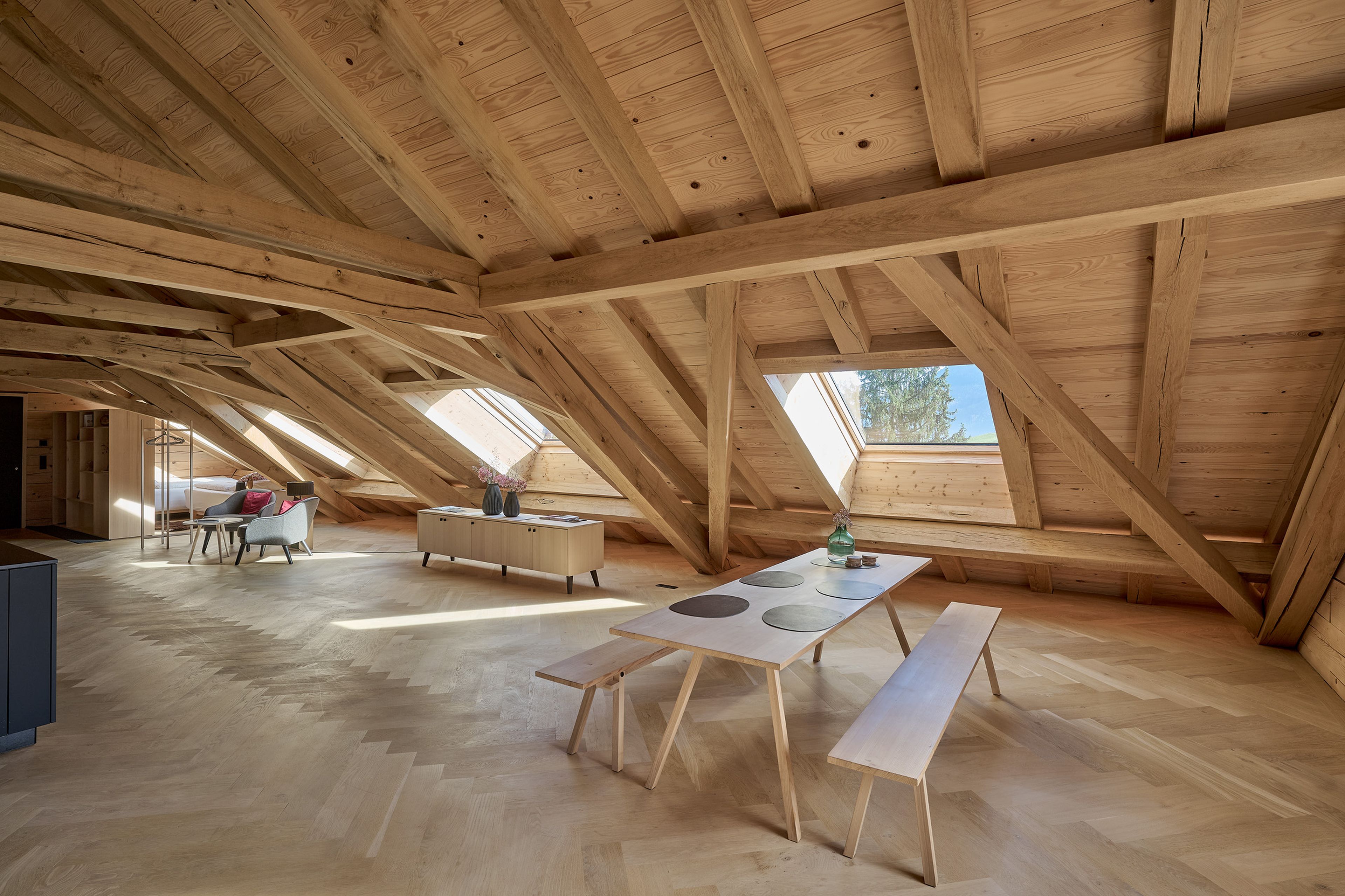 Ein geräumiger, hölzerner Dachboden mit sichtbaren Balken, Dachfenstern, einer langen Bank und einem Mann, der auf dem Sofa sitzt. Sonnenlicht strömt durch die Fenster.