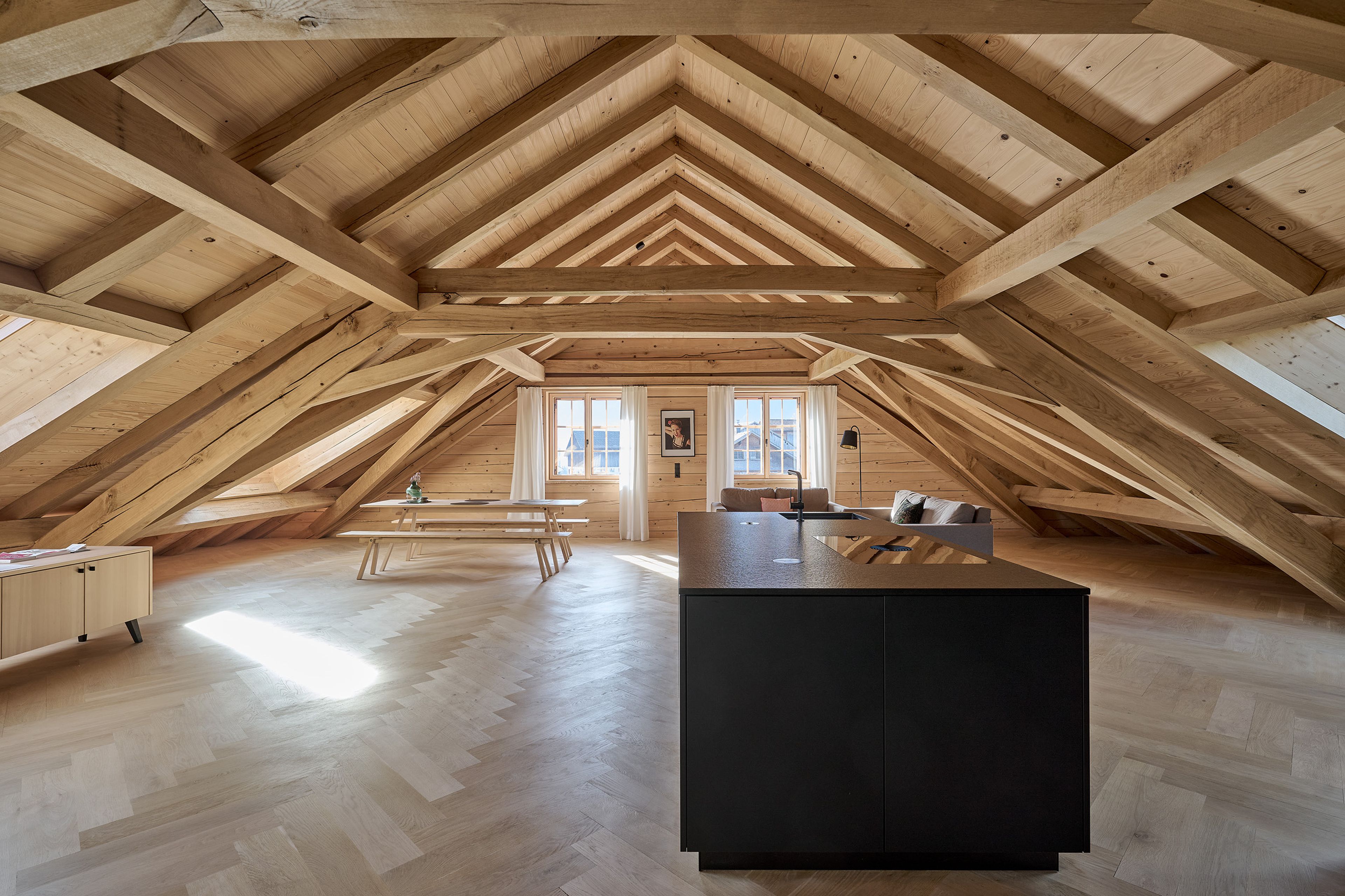 Großzügiges Dachgeschoss mit Holzbalken, moderner Kücheninsel, Esstisch und Sitzecke, mit Oberlichtern und schräger Architektur.