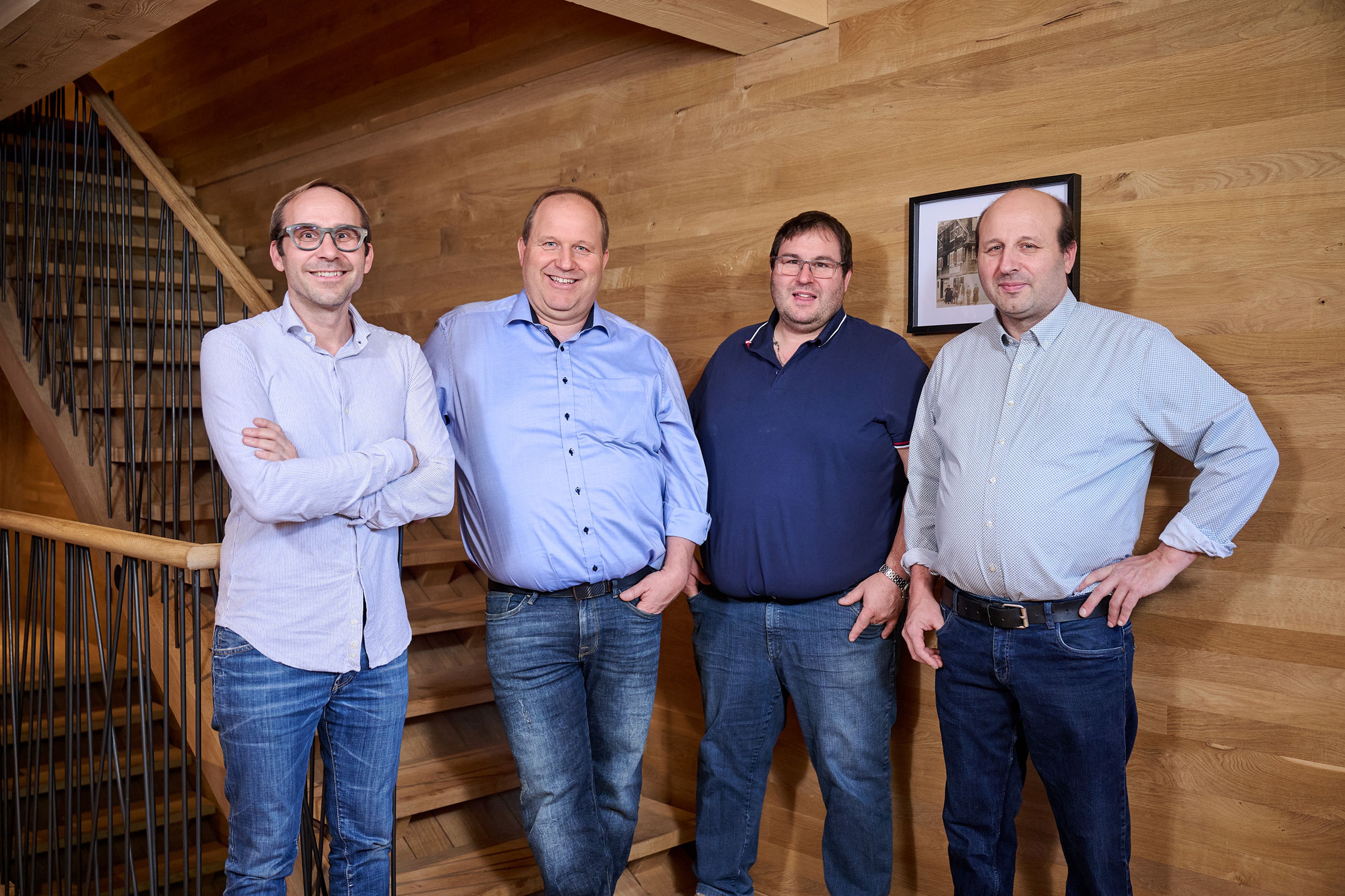 Vier Männer in lässigen Jeans und Hemden stehen lächelnd auf einer Holztreppe.