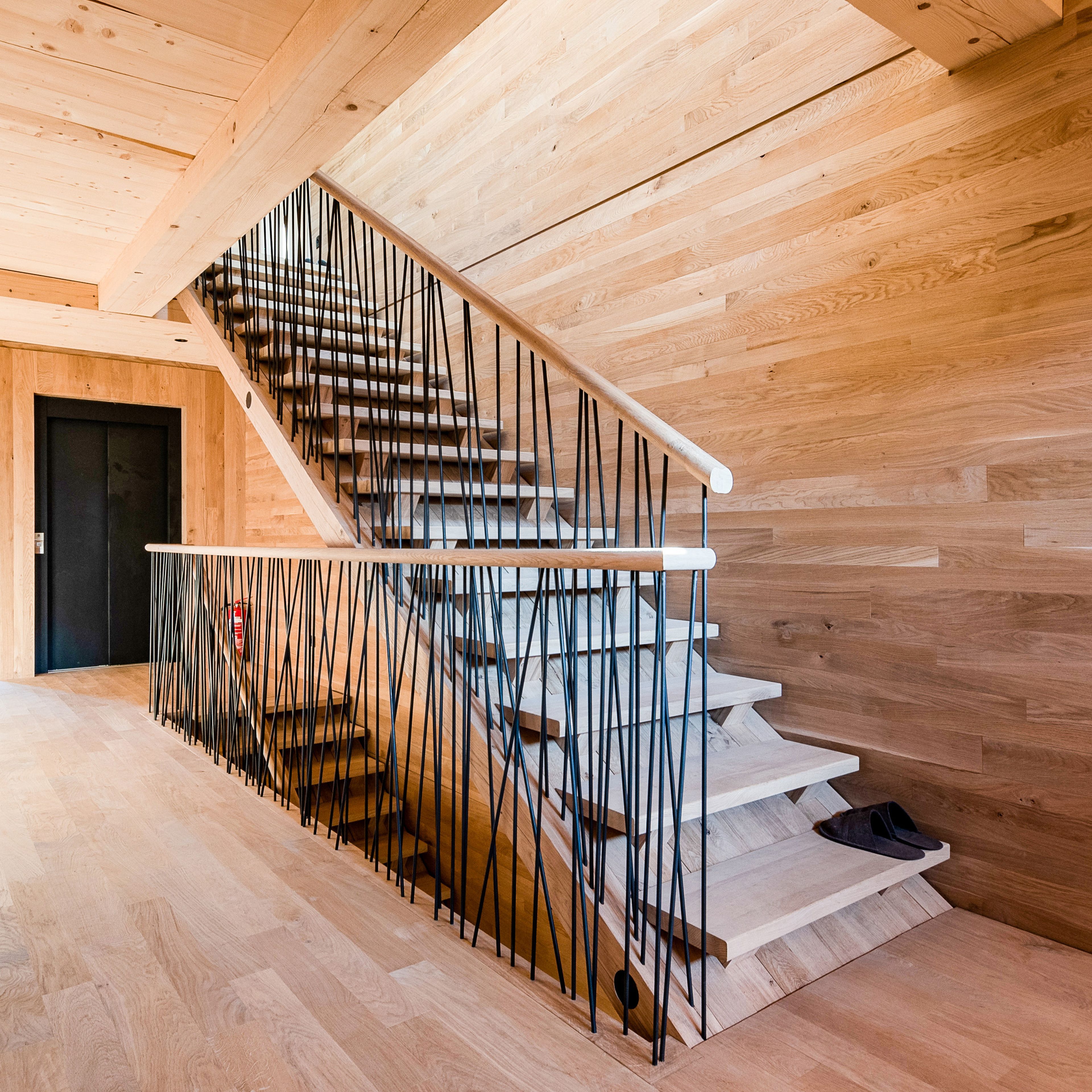 Holztreppe mit modernem Metallgeländer, umgeben von Holzwänden und Holzboden, die zu einem Obergeschoss mit einer dunklen Tür führt.