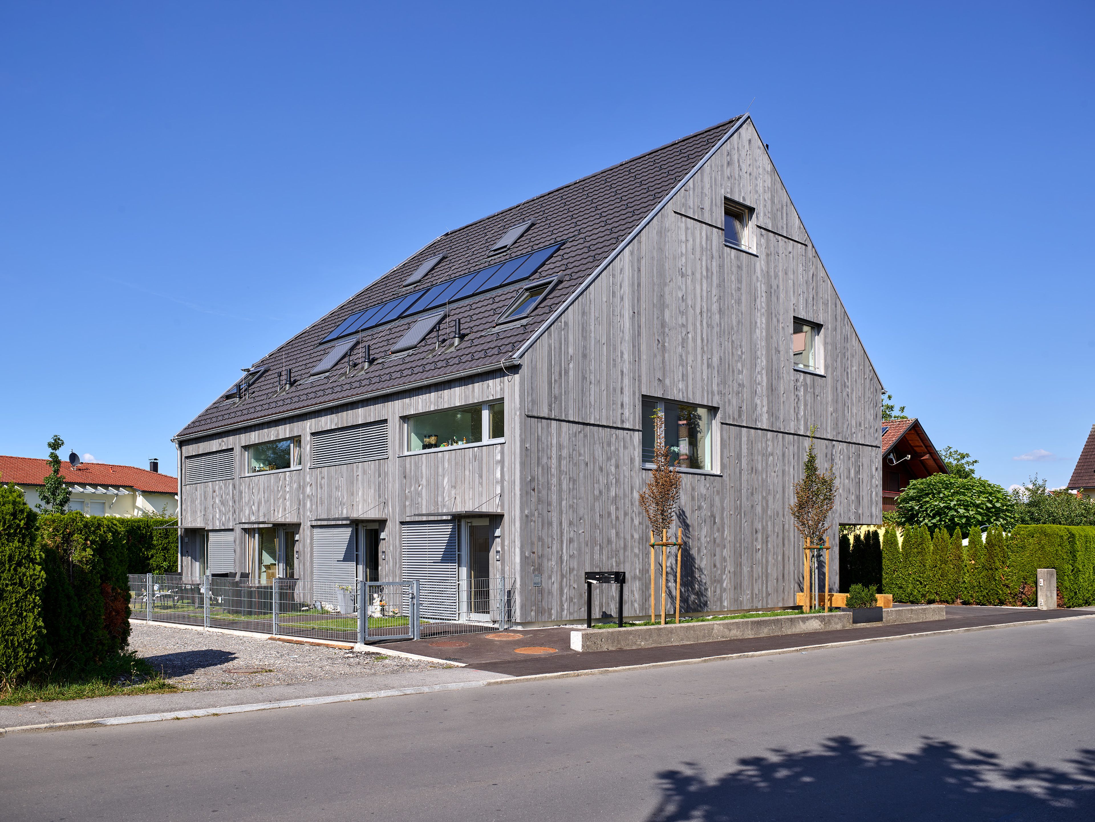 Mehrparteienhaus mit moderner Architektur und grauer Holzfassade; rund um das Gebäude erstreckt sich ein sichtbarer Garten mit Rasenflächen und Bepflanzung.