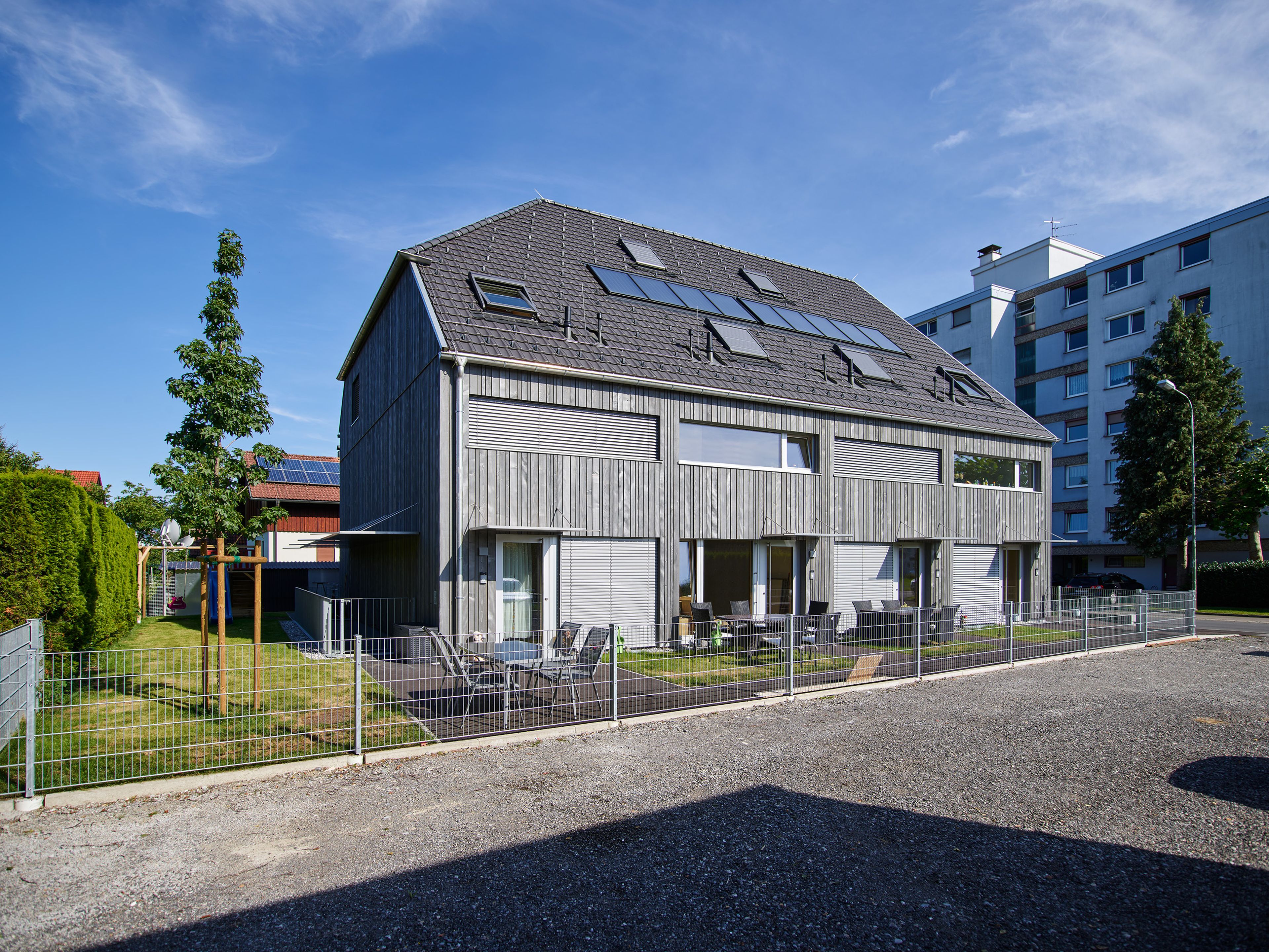 Mehrparteienhaus mit moderner Architektur und grauer Holzfassade; rund um das Gebäude erstreckt sich ein sichtbarer Garten mit Rasenflächen und Bepflanzung.