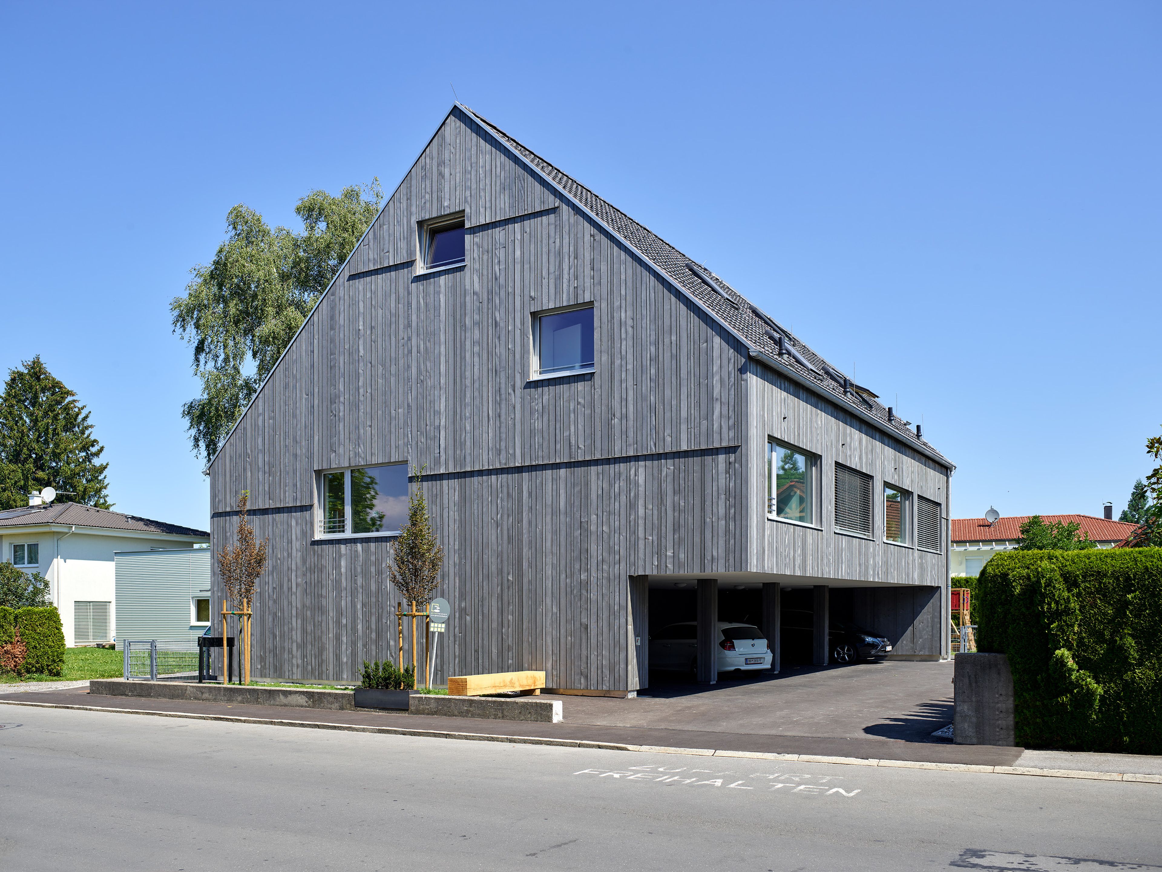 Mehrparteienhaus mit moderner, grauer Holzfassade und Balkonen; rund um das Gebäude erstreckt sich ein sichtbarer Garten mit Rasenflächen und Bepflanzung.