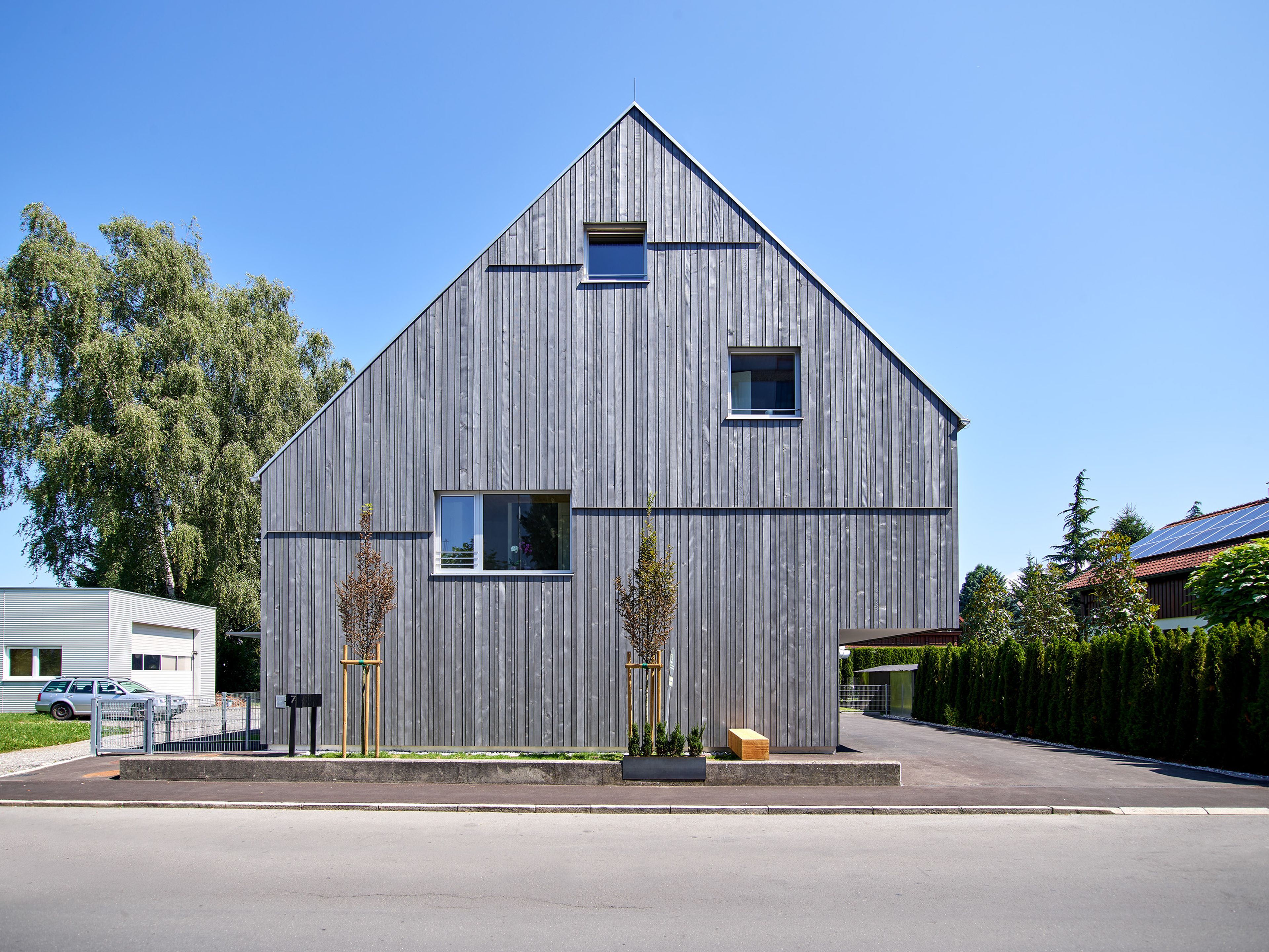 Mehrparteienhaus mit moderner Architektur und grauer Holzfassade; rund um das Gebäude erstreckt sich ein sichtbarer Garten mit Rasenflächen und Bepflanzung.