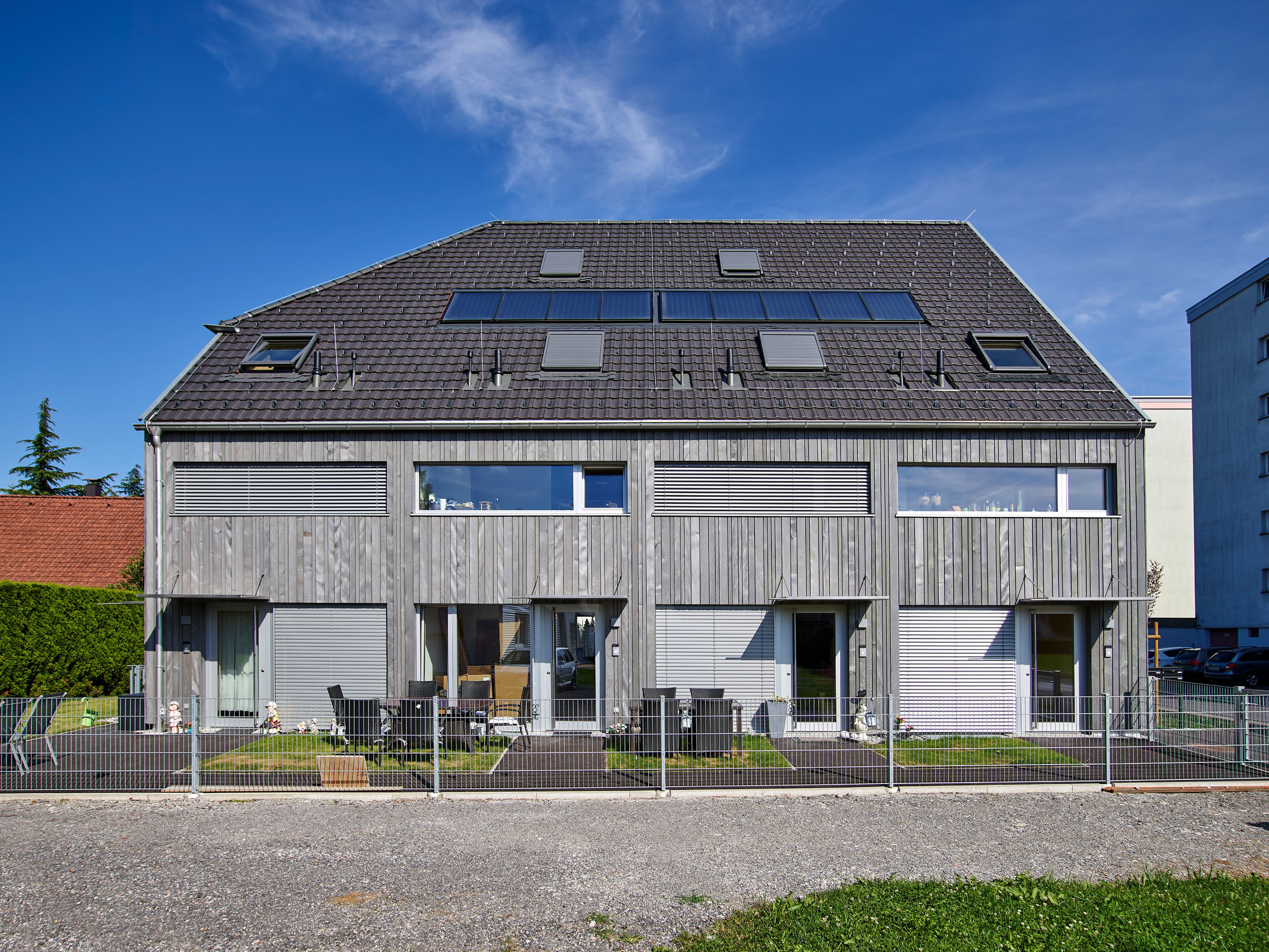 Mehrparteienhaus mit moderner Architektur und grauer Holzfassade; rund um das Gebäude erstreckt sich ein sichtbarer Garten mit Rasenflächen und Bepflanzung.