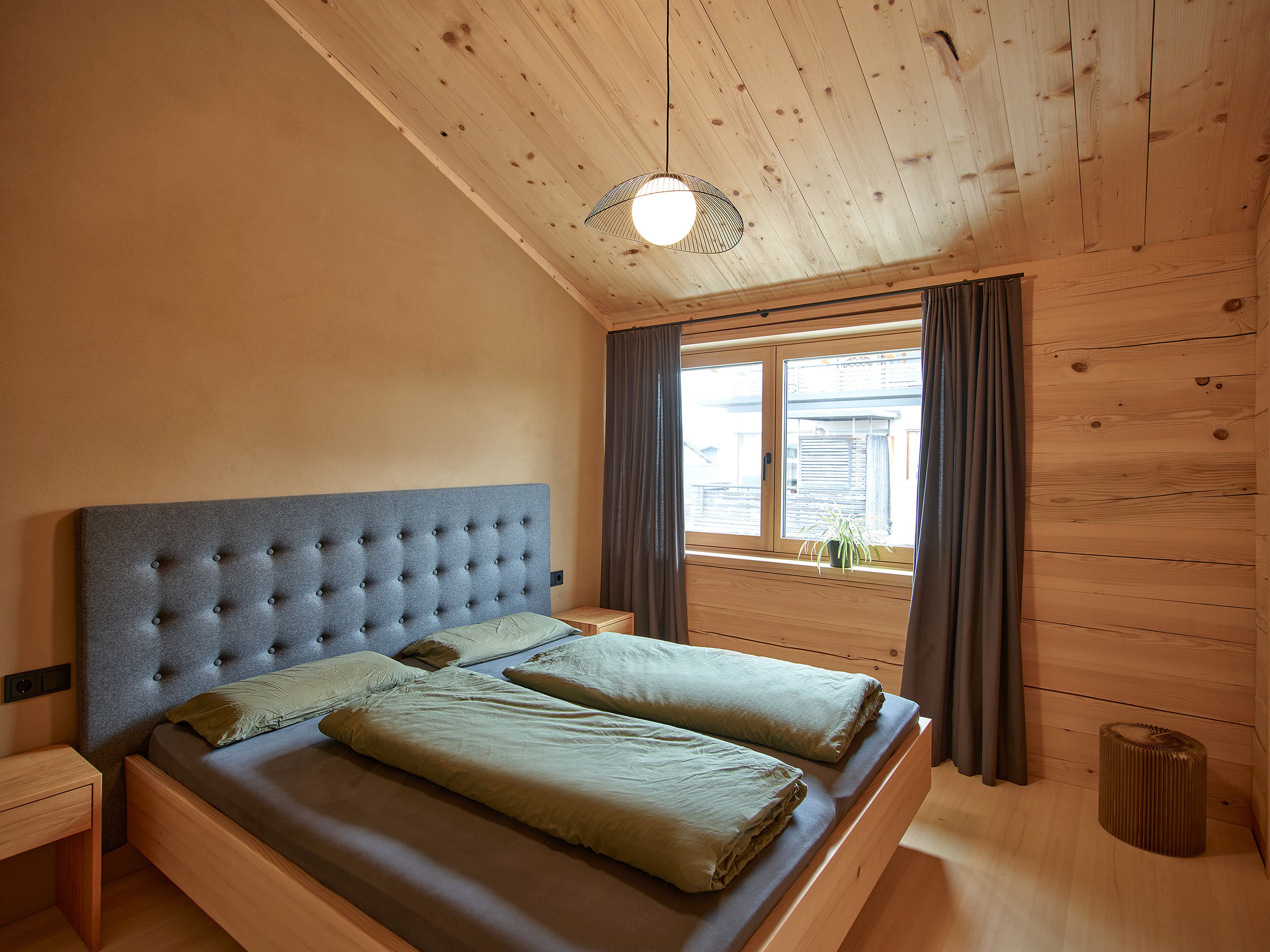 Foto eines Schlafzimmers in einem Holzhaus (Wertvollholzhaus) mit Lehmputzwänden, warmem Licht und dekorativen Vorhängen, die eine gemütliche, natürliche Atmosphäre schaffen.