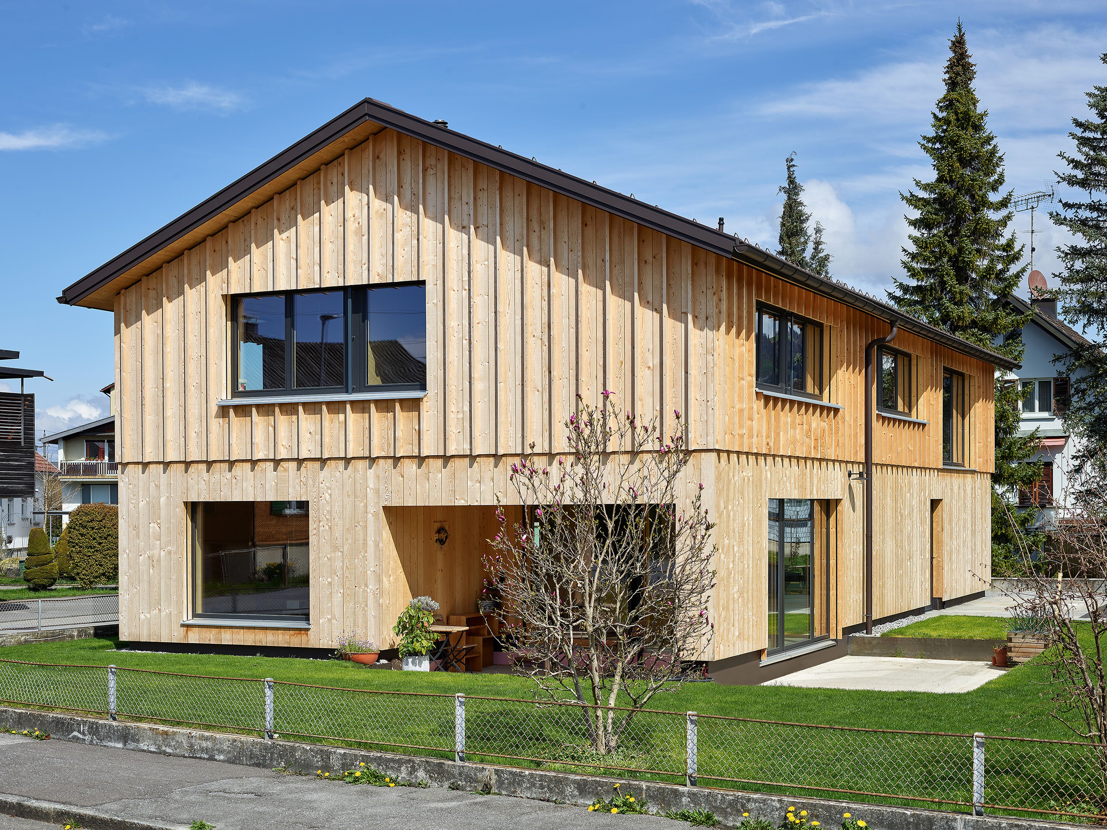 Außenansicht eines Holzhauses in gestrickter Bauweise mit vertikaler, stehender Holzfassade, umgeben von einem gepflegten Garten und natürlicher, harmonischer Umgebung.