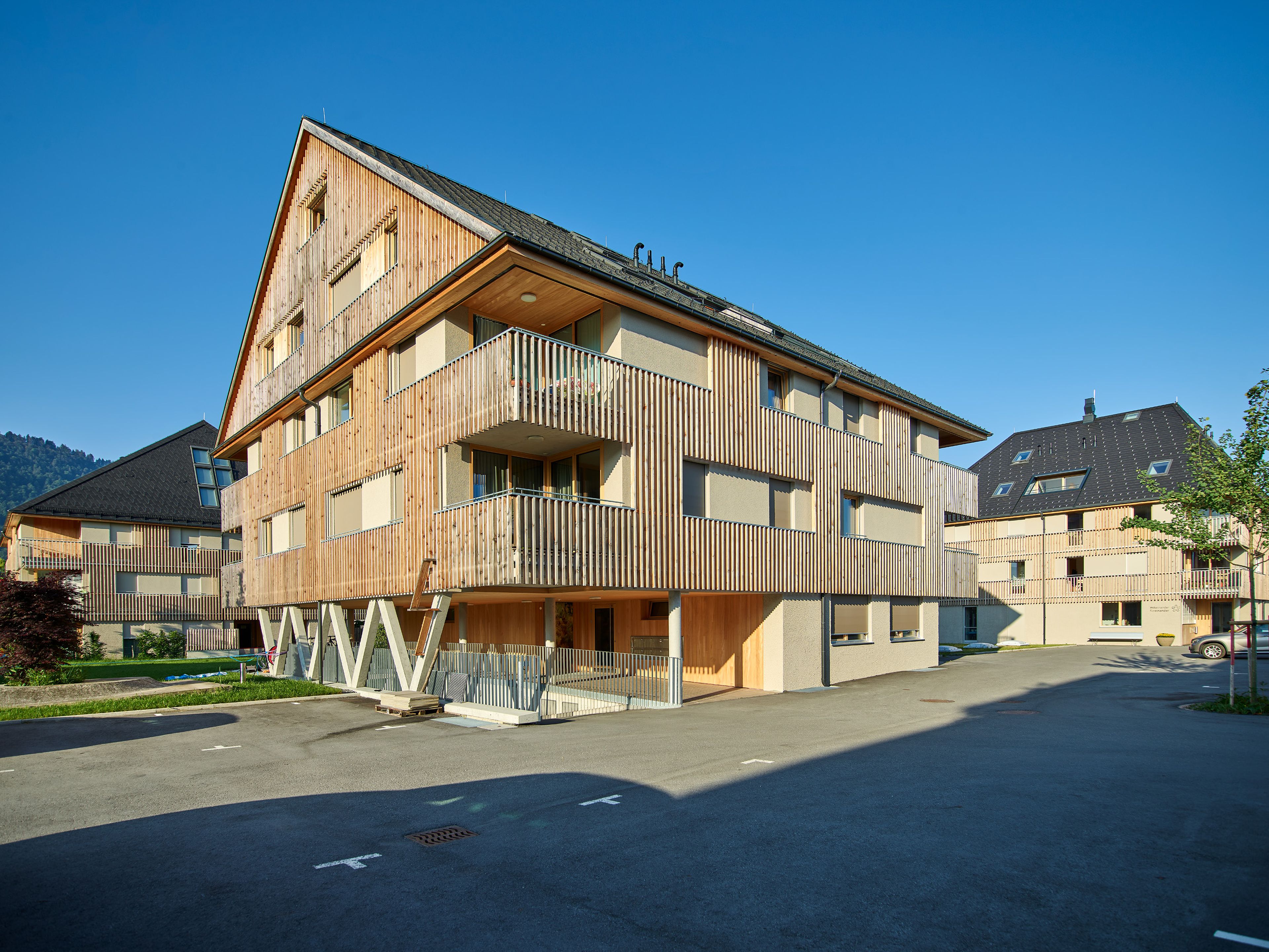 Wohnanlage in Holzbauweise mit moderner Architektur und Holzfassade und Satteldach; rund um das Gebäude erstreckt sich viel Grünfläche.