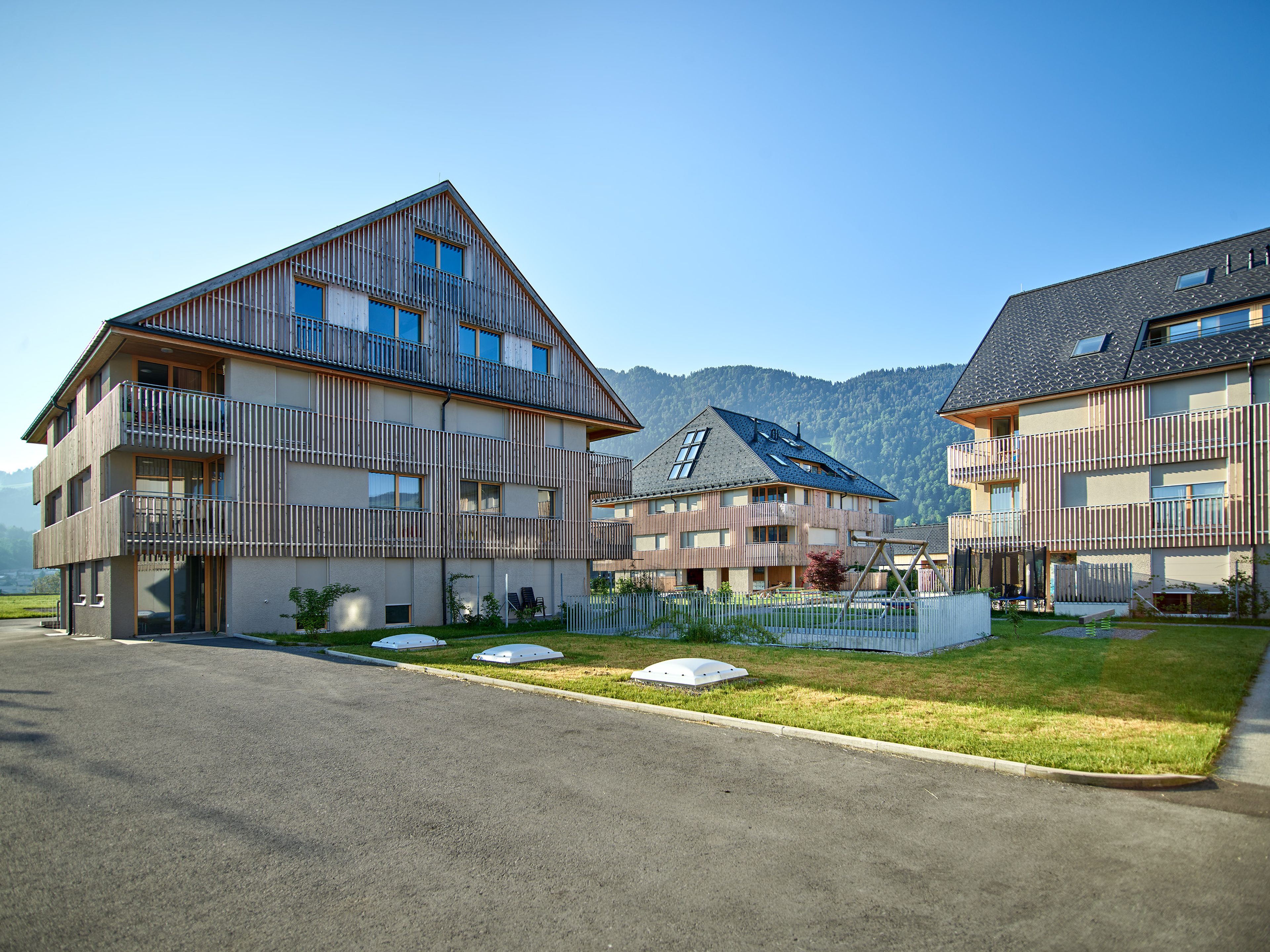 Wohnanlage in Holzbauweise mit moderner Architektur und Holzfassade und Satteldach; rund um das Gebäude erstreckt sich viel Grünfläche.