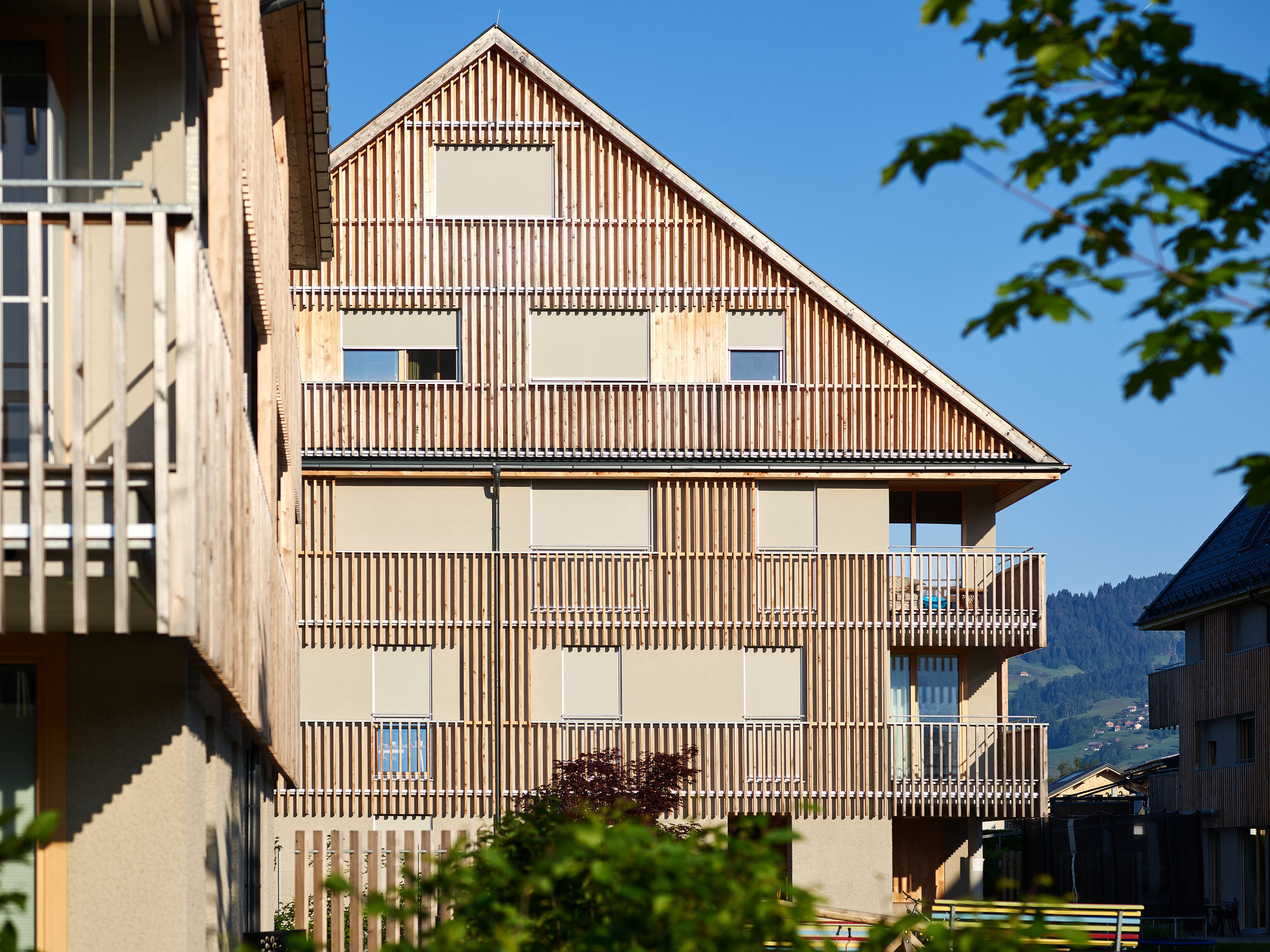 Wohnanlage in Holzbauweise mit moderner Architektur und Holzfassade und Satteldach; rund um das Gebäude erstreckt sich viel Grünfläche.