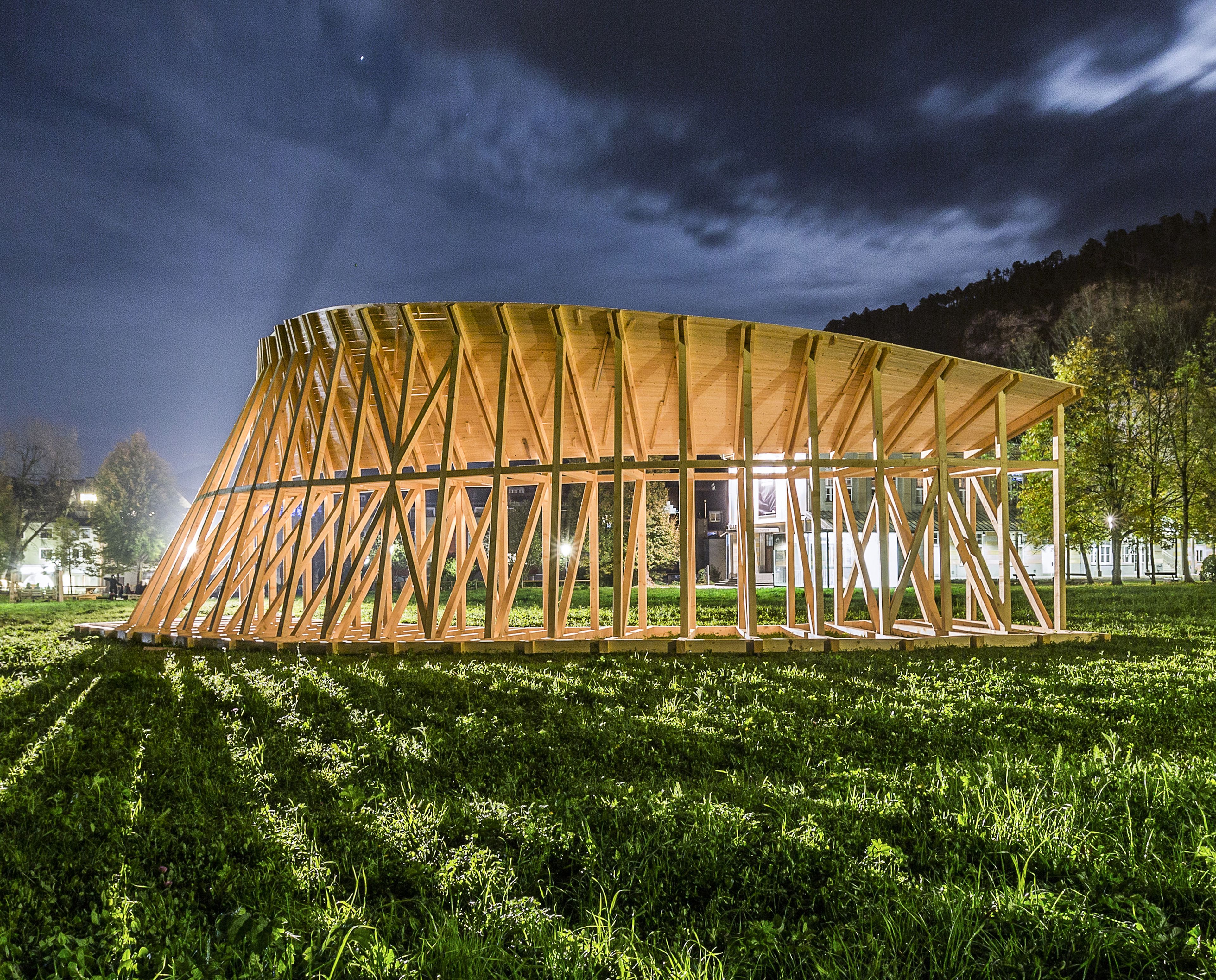 Auf einem grasbewachsenen Feld, unter einem bewölkten Himmel, wird nachts ein moderner Holzpavillon als Kunstwerk/Installation mit geschwungenem Dach beleuchtet.