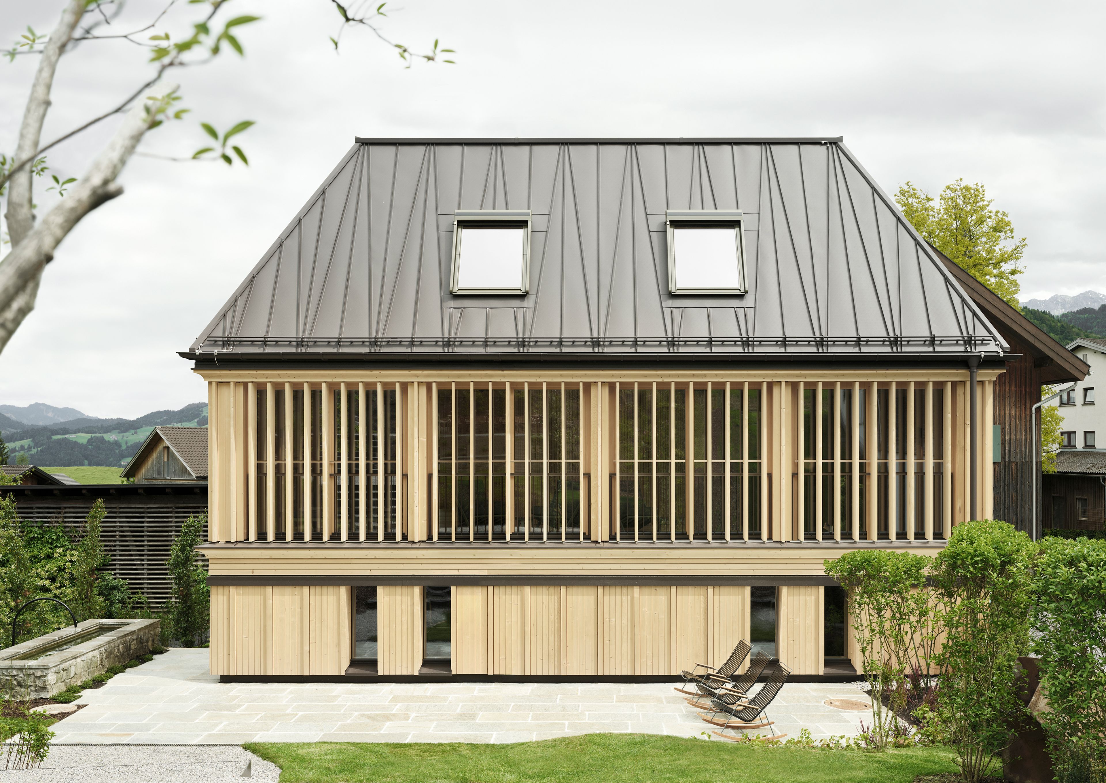 Modernes Holzhaus mit Metalldach, vertikalen Holzlamellen im Obergeschoss und zwei Dachfenstern. Steinterrasse mit zwei Liegestühlen davor.