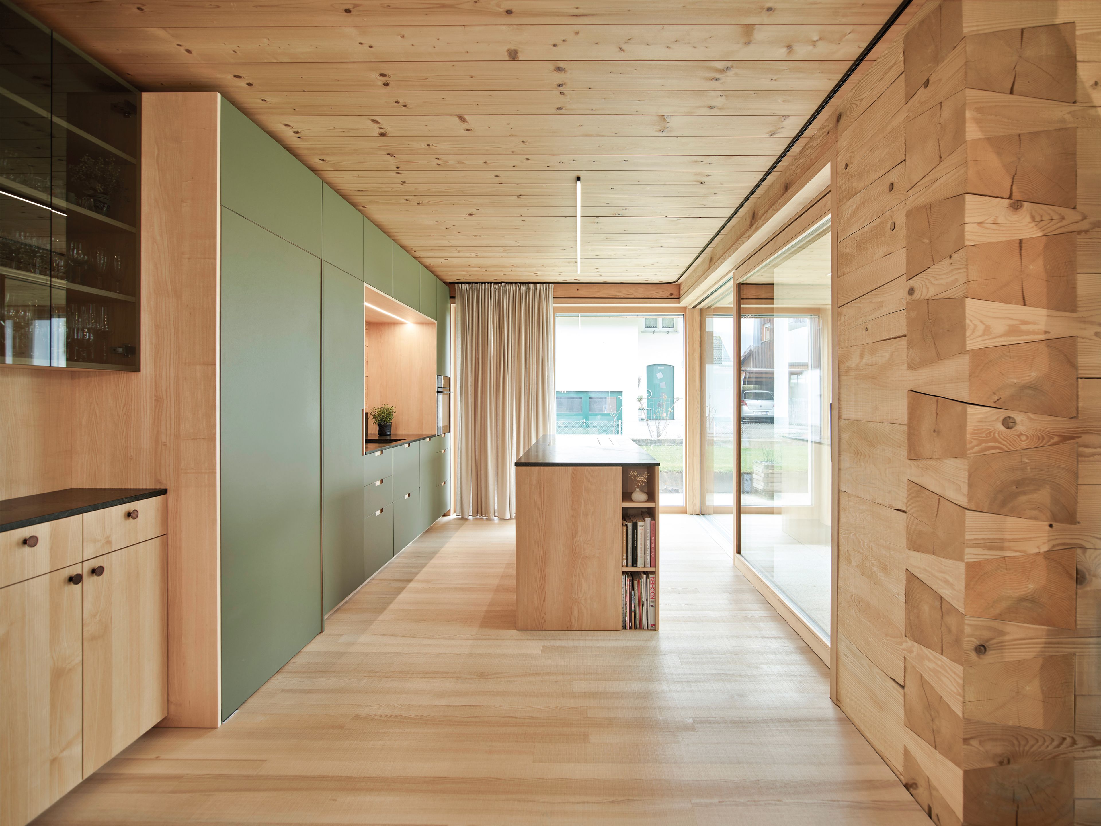 Moderne Küche in einem Holzhaus mit hellen Holz- und olivfarbener Küche, einer Kochinsel mit Bücherregalen, großen Fenstern und einer Holzdecke und Holzboden.