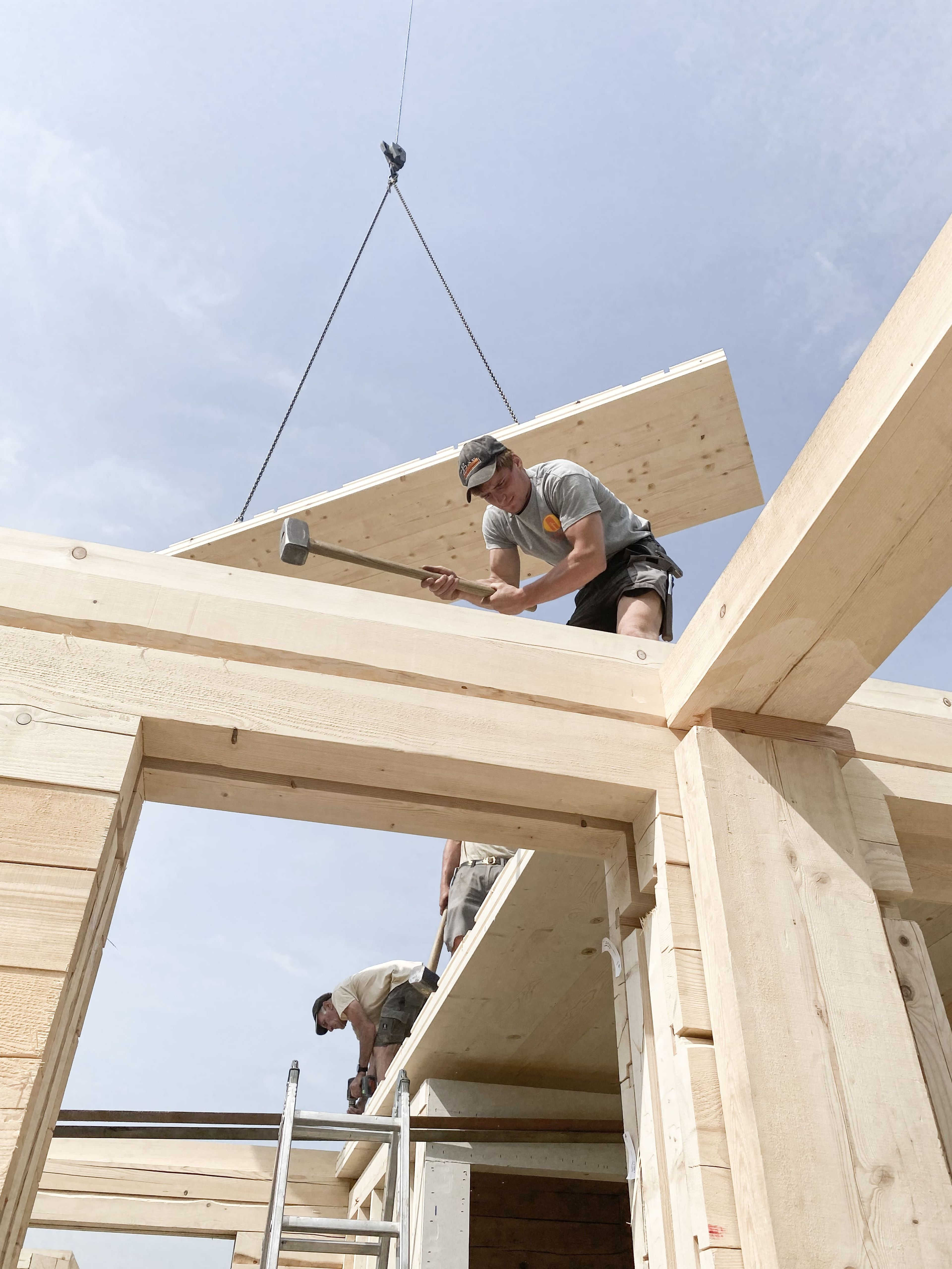 Zwei Bauarbeiter montieren eine Holzkonstruktion; einer steht auf einer Leiter, der andere hämmert von oben, während ein Kran eine Holzplatte anhebt.