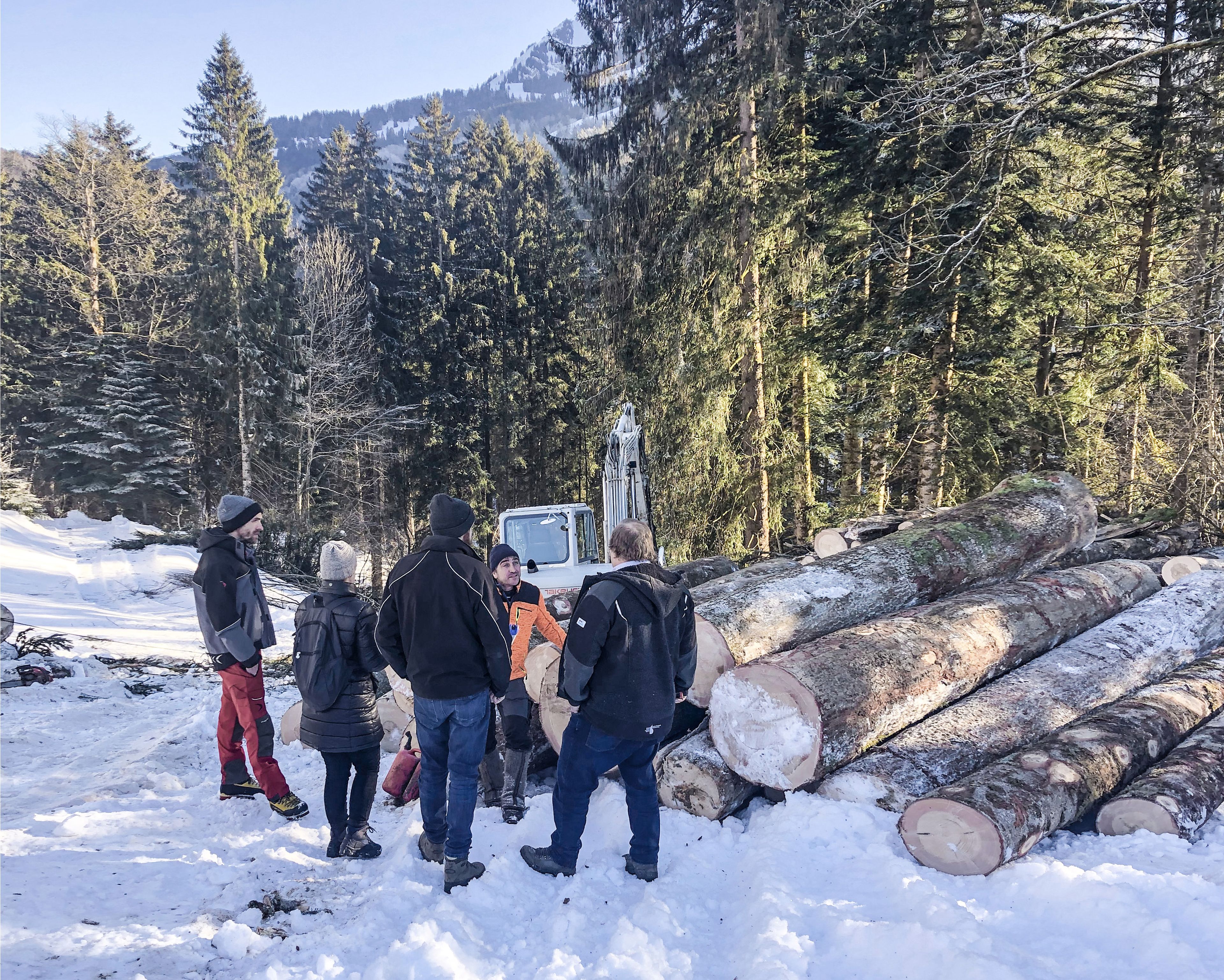 A group of people stands in a snowy forest next to large cut logs and a small excavator, having a discussion.