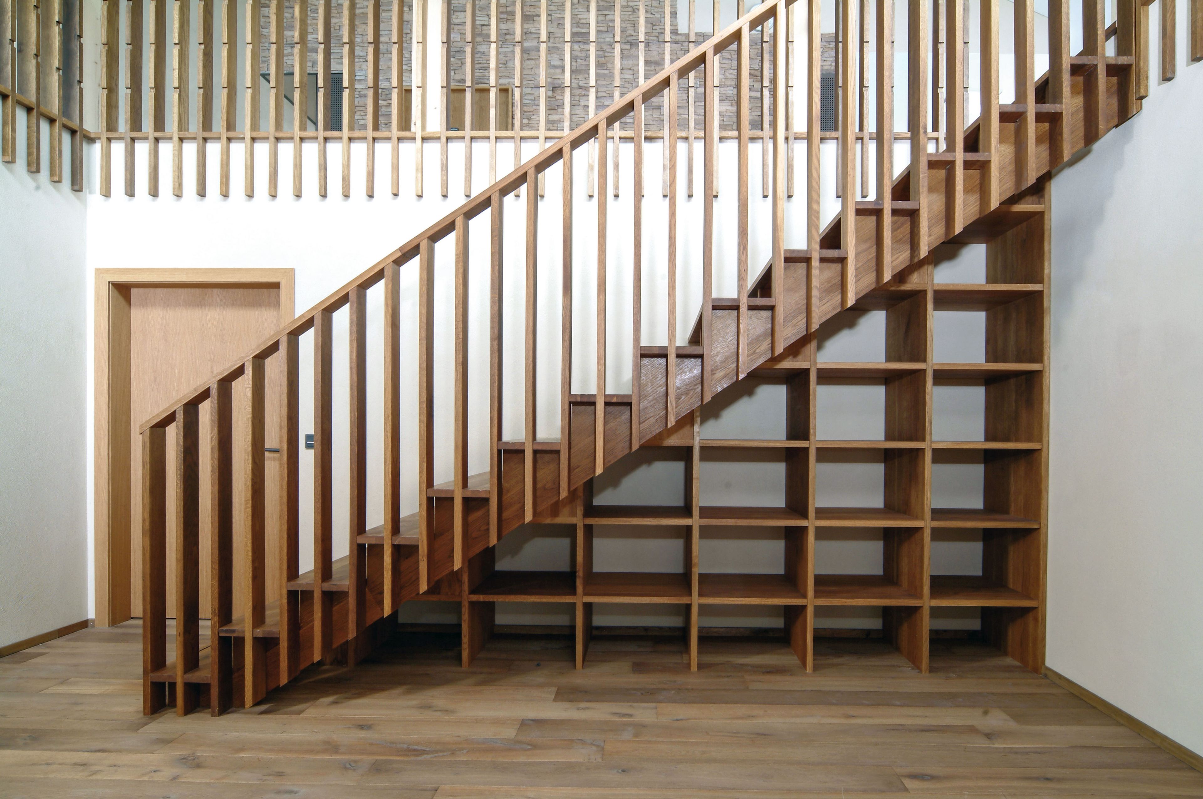 Eine Holztreppe mit senkrechten Geländern und integrierten offenen Regalen darunter, vor einer weißen Wand und einem holzgetäfelten Boden.