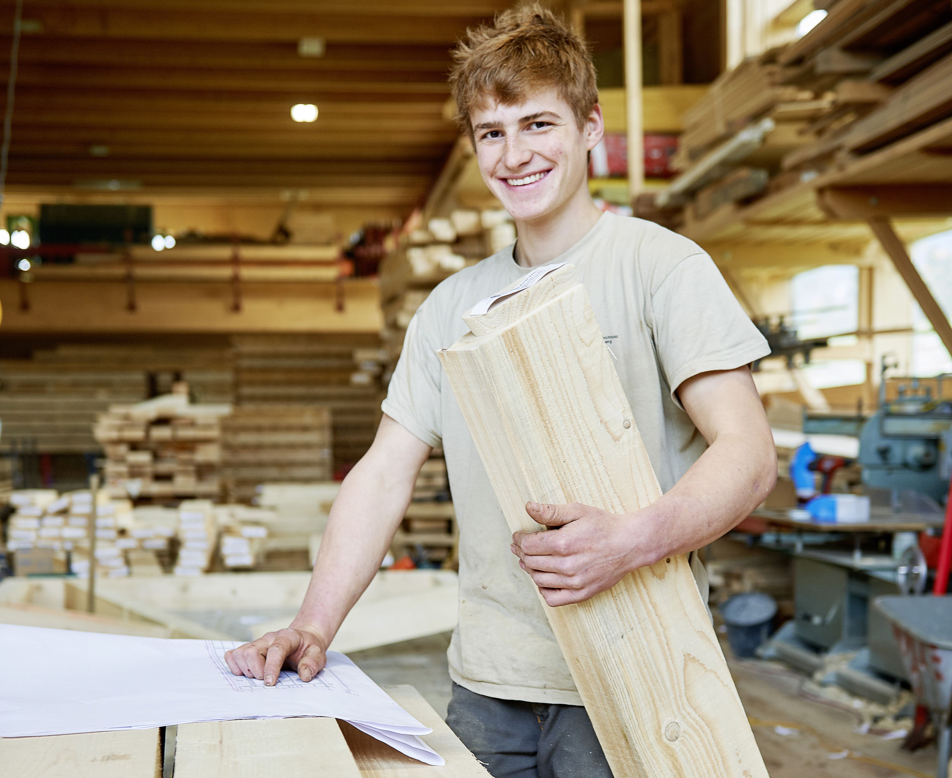 Ein lächelnder junger Mann in einer Werkstatt hält ein Holzbrett in der Hand; auf dem Tisch liegen Pläne. Umgeben von Holz und Werkzeugen in einem hellen Raum.