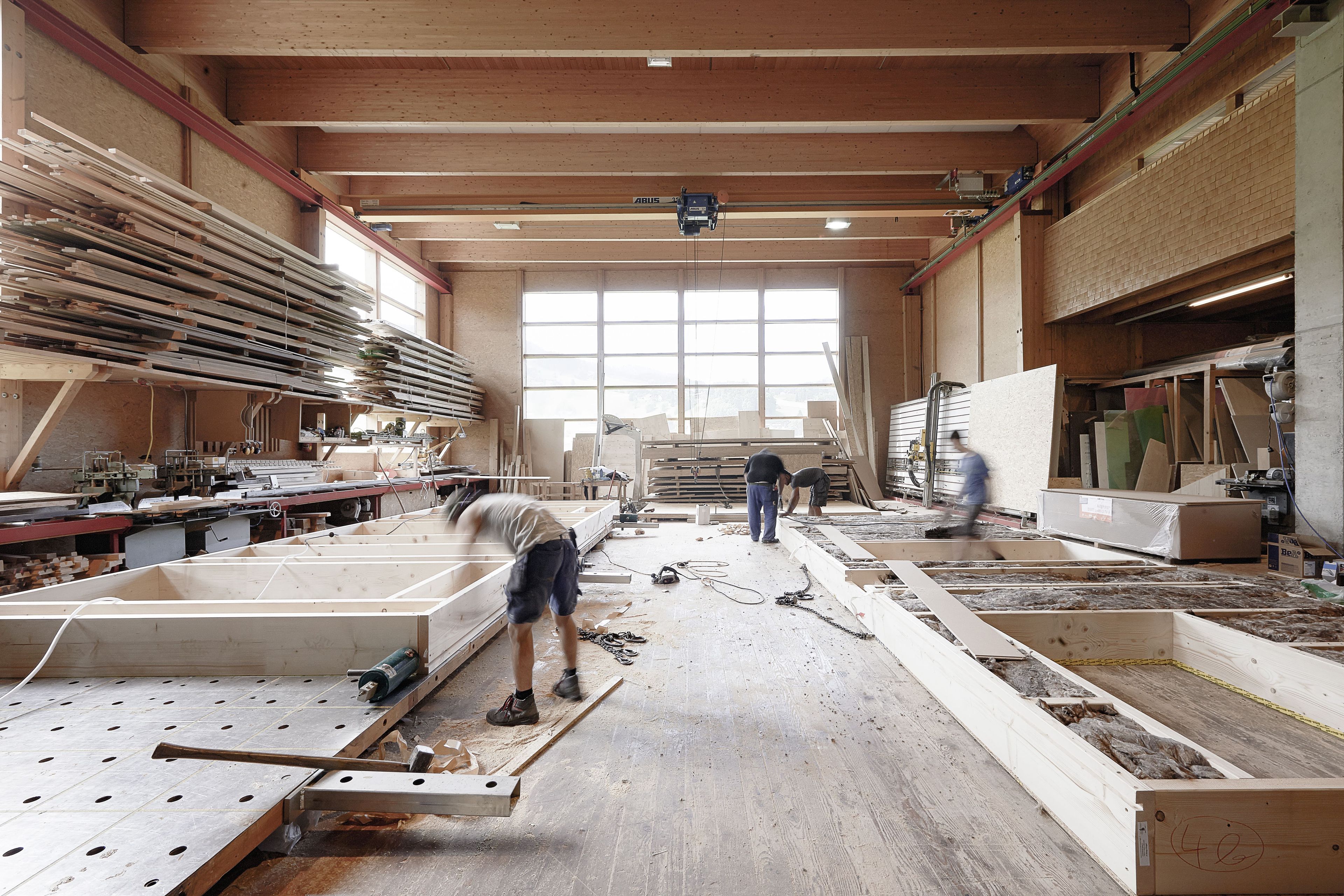 Arbeiter in einer geräumigen Holzwerkstatt, die große Holzrahmen bauen, umgeben von Holzstapeln und verschiedenen Werkzeugen.