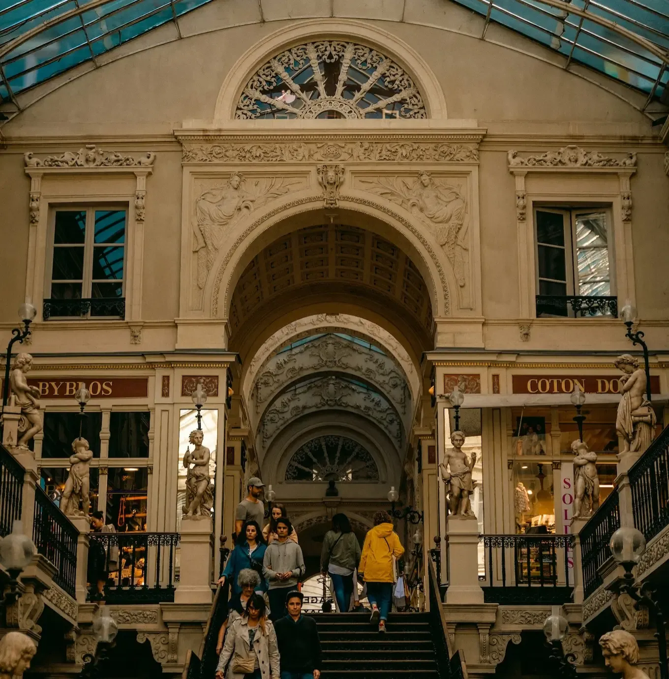 Des personnes descendant un grand escalier dans une galerie marchande historique, ornée de détails architecturaux raffinés, de statues et d'une verrière au-dessus.