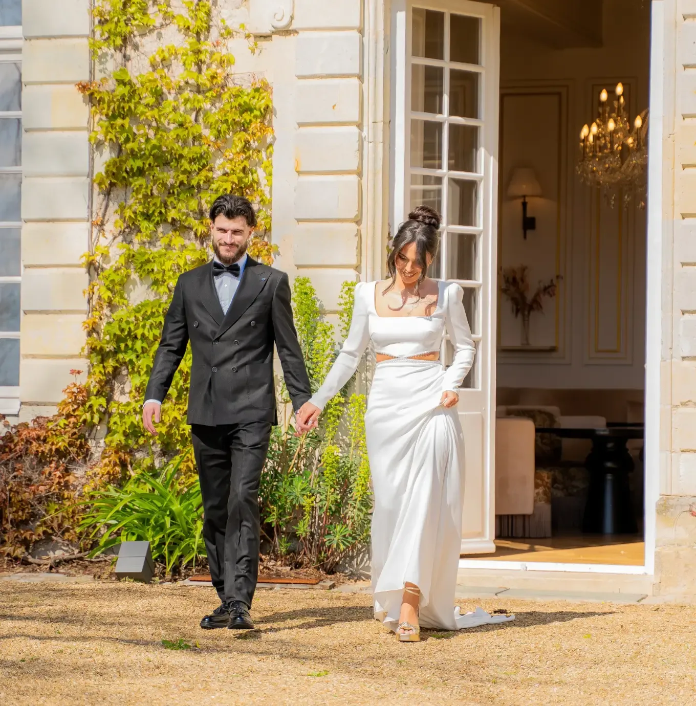 Un couple en tenue élégante marche main dans la main devant un immeuble bien éclairé, dont l'entrée est bordée de vignes et de plantes vertes.