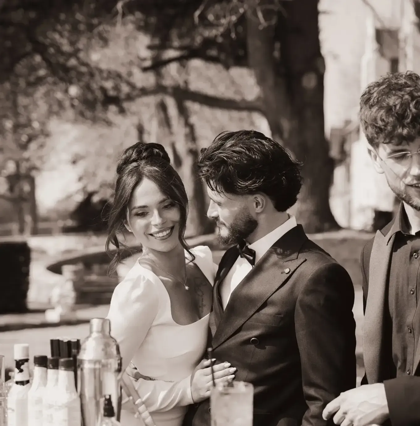 Un couple en tenue de soirée sourit près d'un bar en plein air, où l'on aperçoit des bouteilles et des shakers ; une autre personne se tient debout à droite, vêtue d'une chemise noire.