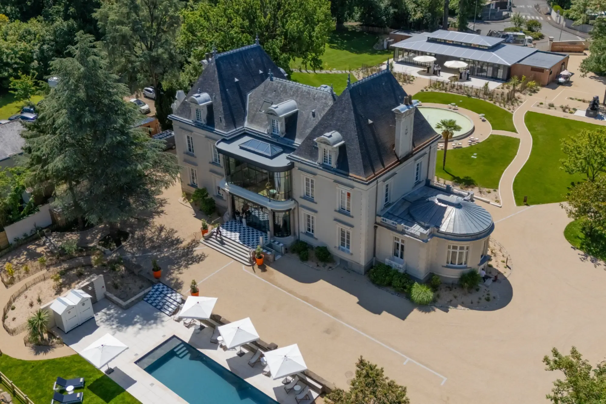 Vue aérienne du Château du Portereau élégant au toit sombre, entouré de jardins paysagers, d'une allée circulaire et d'une piscine agrémentée de parasols blancs.
