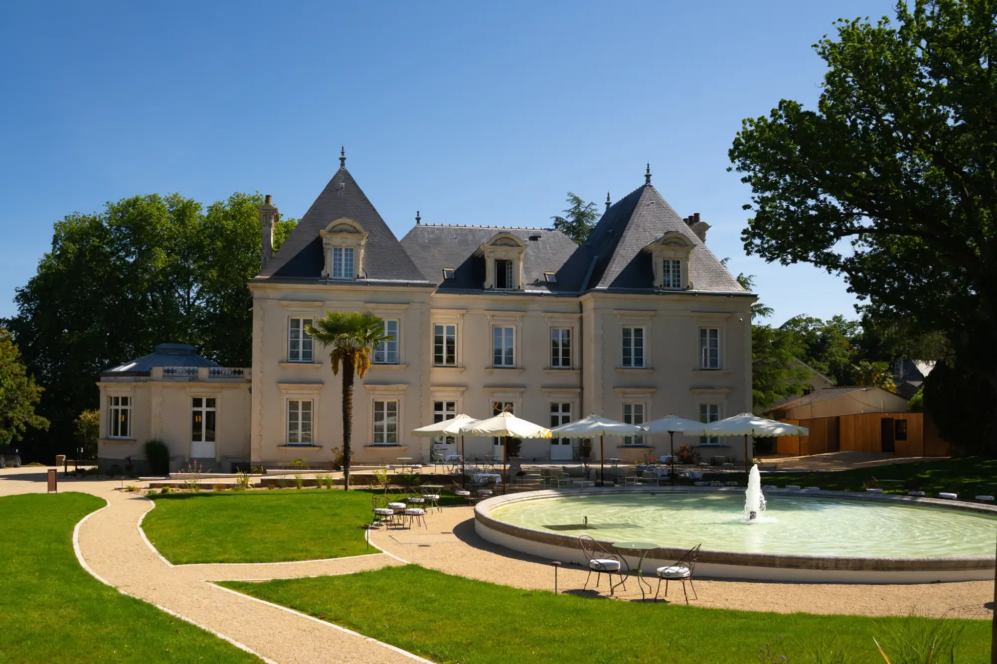 L'élégante façade du Château du Portereau avec de hauts toits en ardoise, des parasols blancs, une terrasse et une fontaine circulaire, le tout sous un ciel bleu.