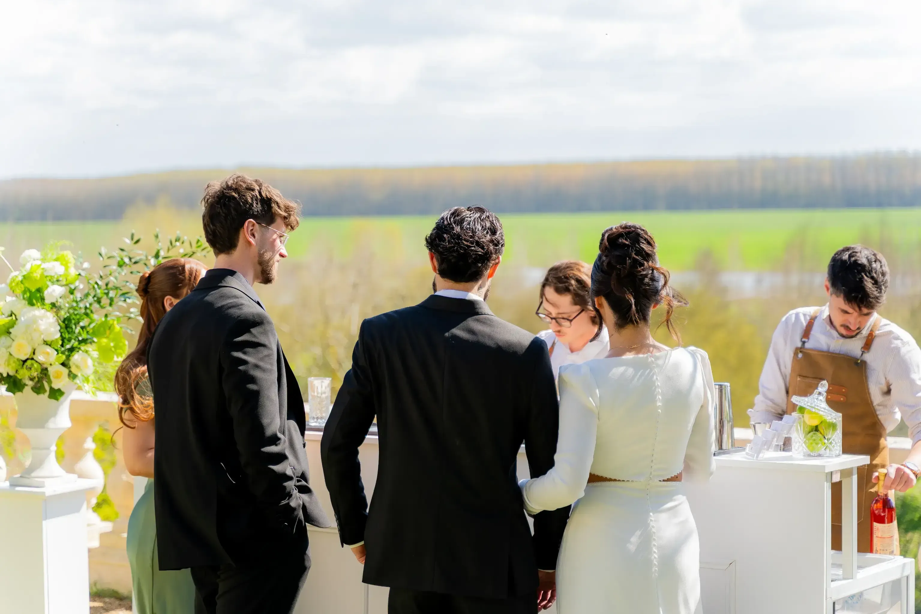 Des personnes en tenue de mariés se rassemblent autour d'un bar en plein air, avec une pelouse verte en arrière-plan et des barmans qui servent des boissons.