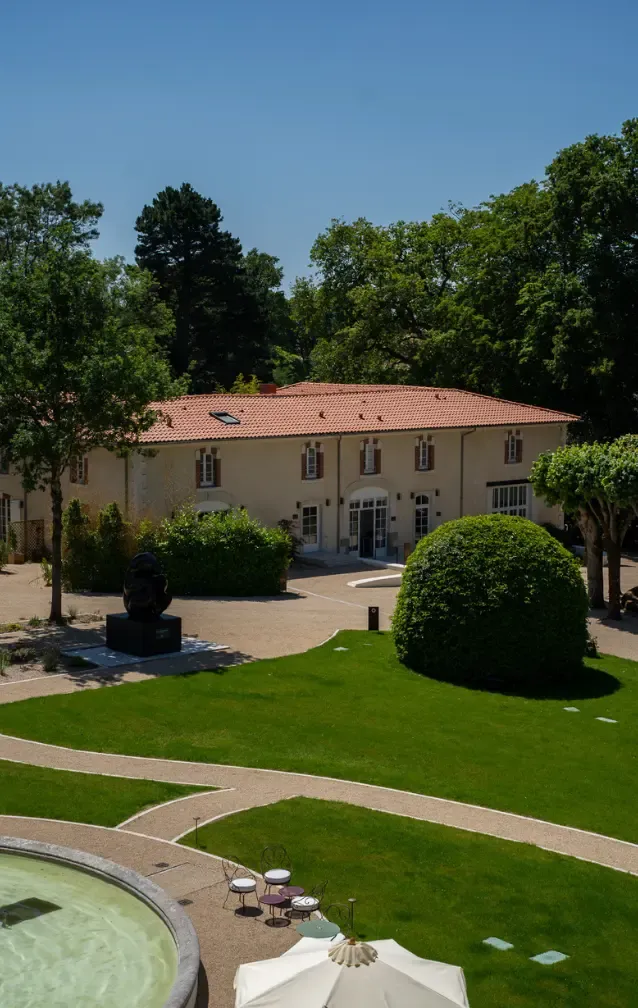 Un jardin verdoyant agrémenté d'une sculpture, de sentiers sinueux, d'une terrasse avec des sièges, et du pavillon du Château entouré de grands arbres.