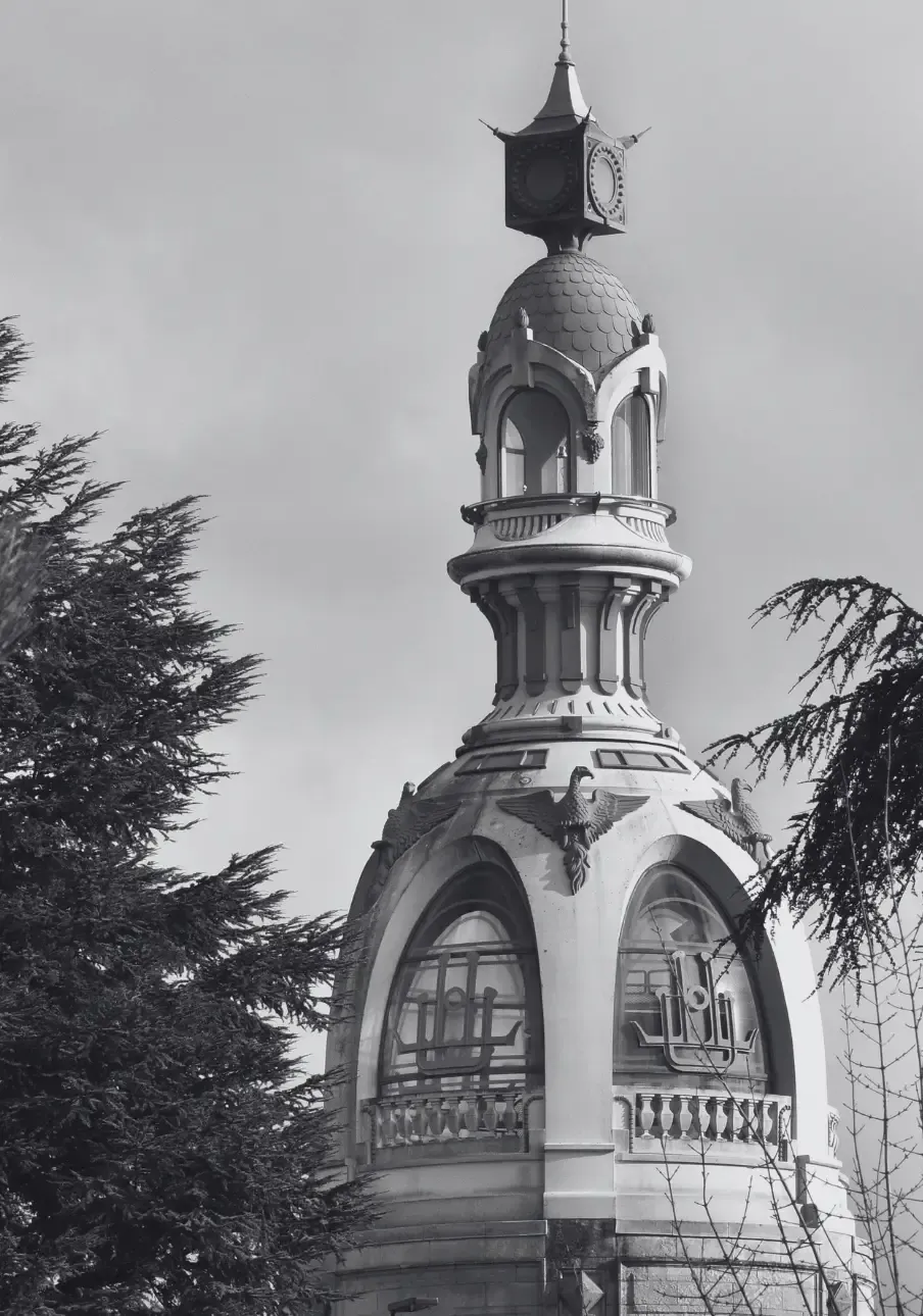 Photo en noir et blanc de la tour LU richement décorée, avec des fenêtres cintrées et des motifs complexes, encadrée par des branches d'arbres.