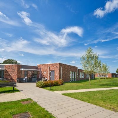 Low brick community buildings with paved paths, benches, young trees and green lawn under a bright blue sky.